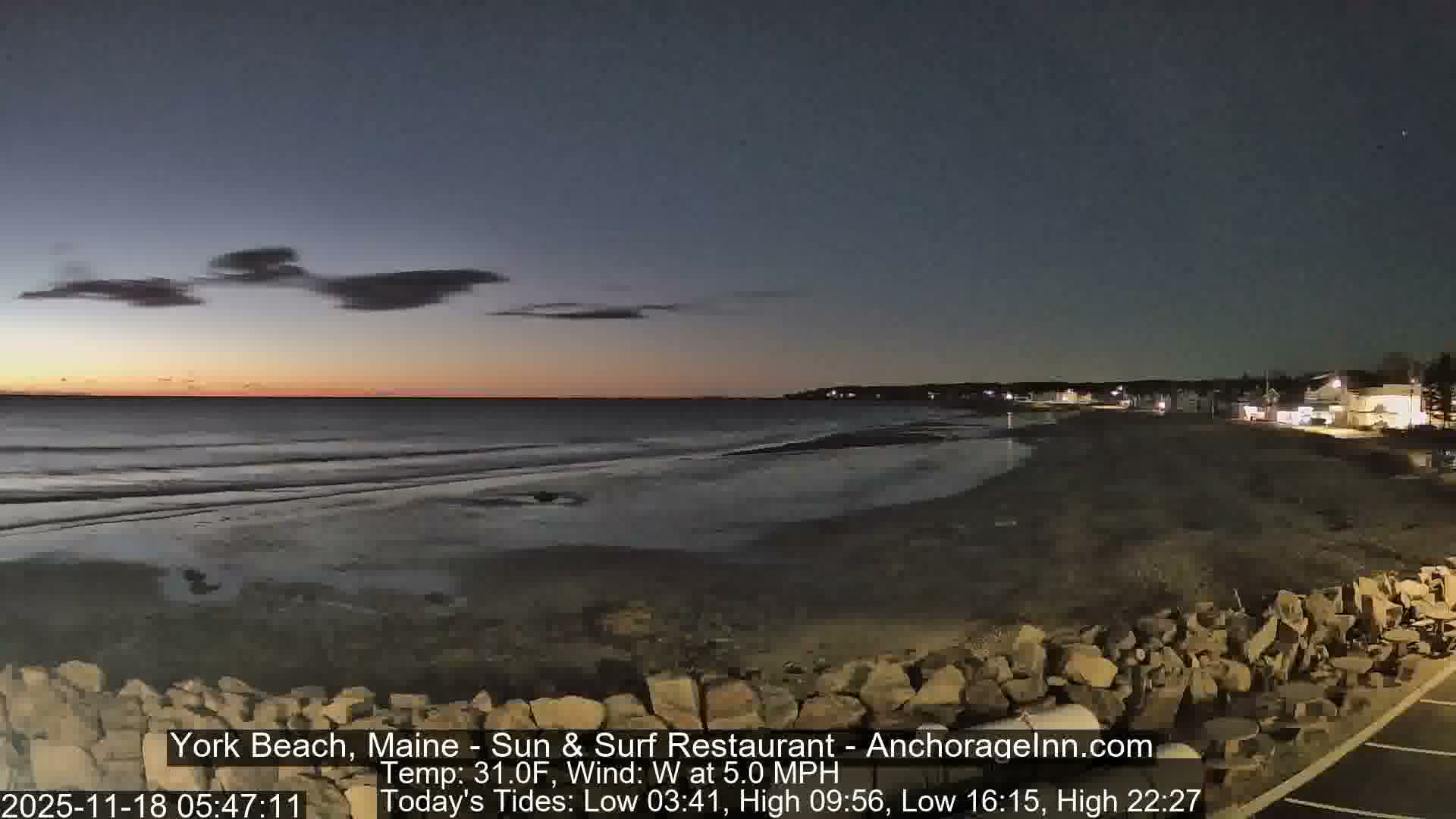 A dark, cloudy night reveals waves breaking on a beach with a rocky foreground and distant illuminated buildings along the coastline.