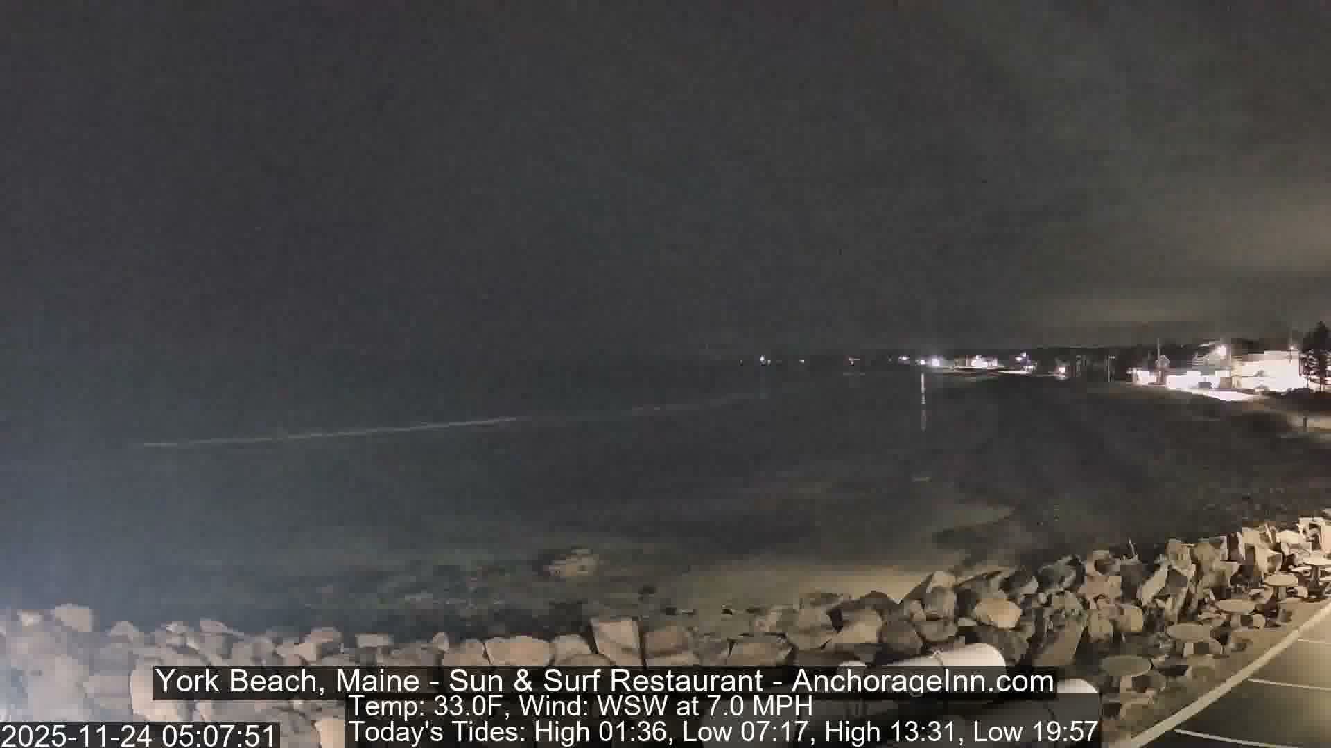 An overcast night scene reveals a dark, rocky beach stretching along calm ocean waters, with illuminated coastal buildings visible in the distance and an empty patio with tables in the foreground.