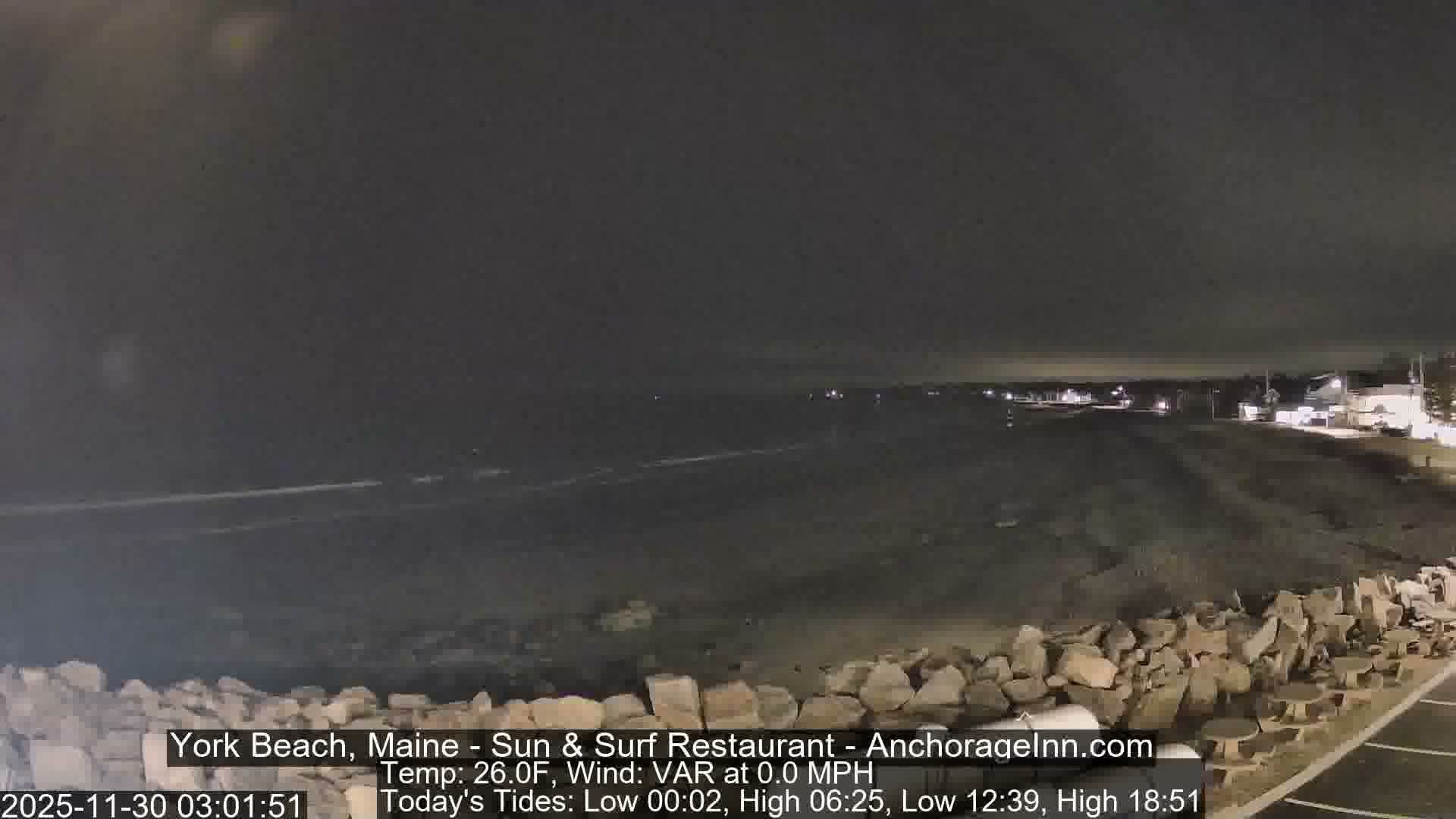 A nighttime view reveals a dark beach with gentle waves under an overcast sky, with scattered lights from coastal buildings illuminating the horizon and rocks in the foreground.