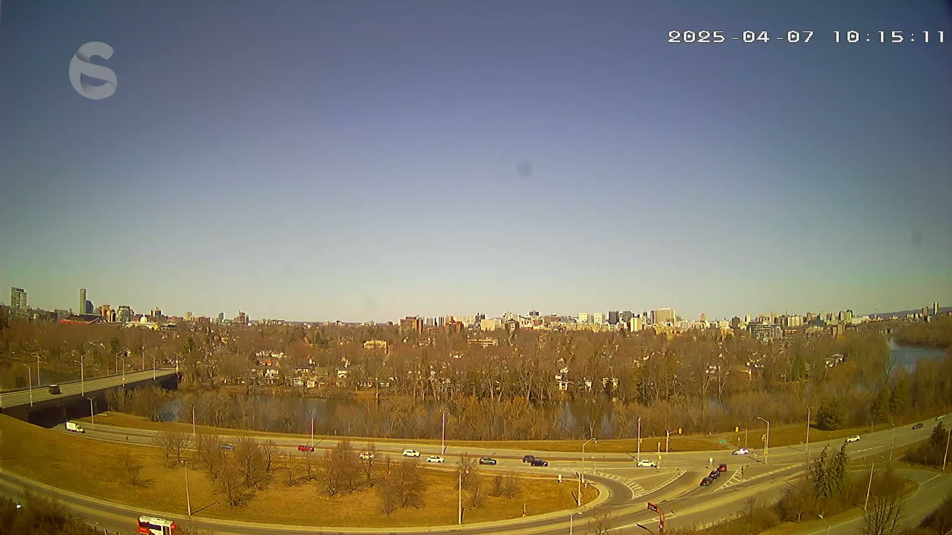 A clear, sunny day shows a highway curving around a river, with a city skyline visible in the distance and sparse early-spring vegetation along the roadside.