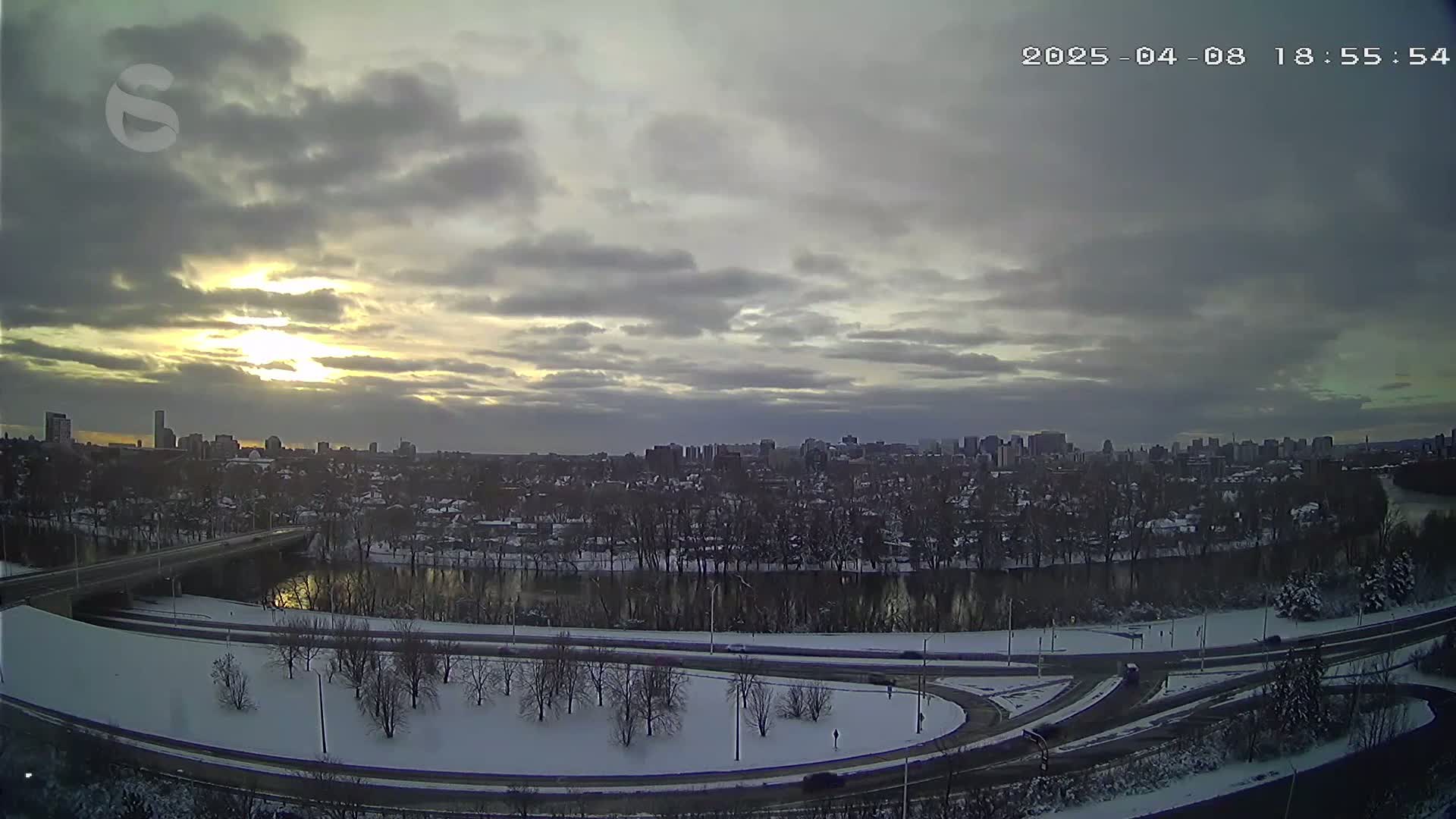 A snow-covered city skyline is visible at sunset under a cloudy sky, with a river and highway curving through a snowy landscape in the foreground.