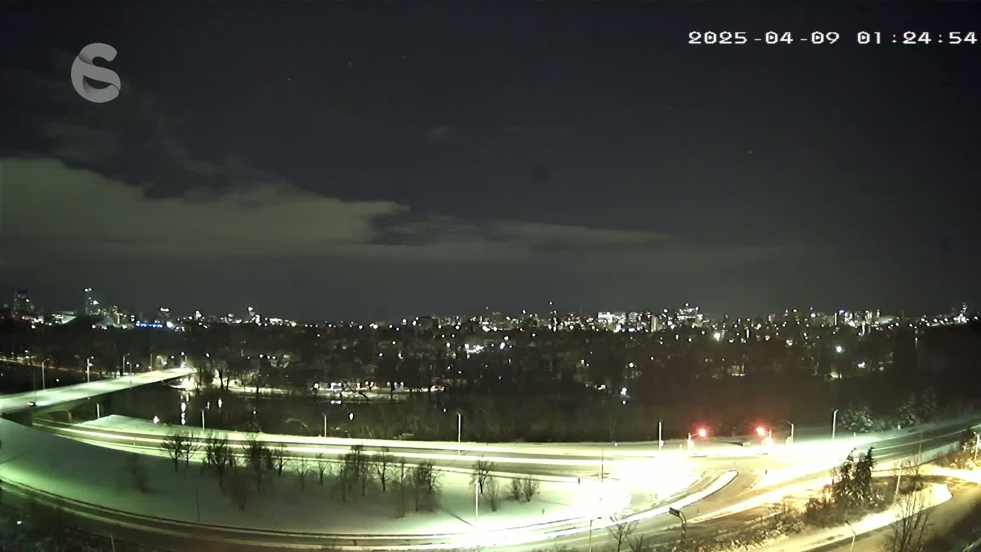A snow-covered highway interchange curves around a river, leading to a city skyline under a partly cloudy night sky.