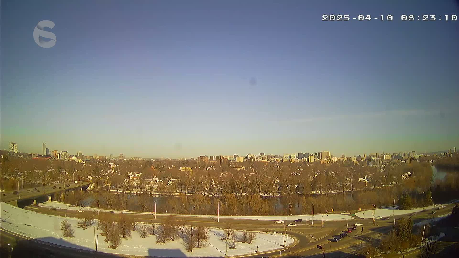 A clear, sunny day reveals a cityscape across a river, with a roadway curving around a snow-dusted park in the foreground.
