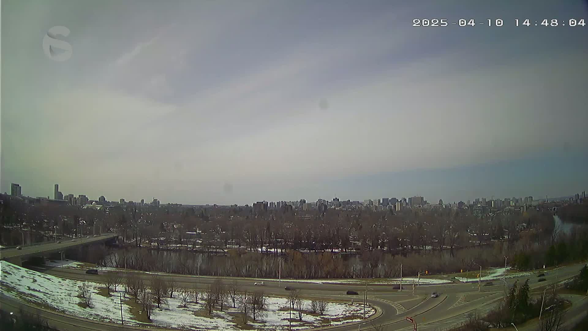 A mostly cloudy day reveals a cityscape beyond a river, with bare trees lining the riverbanks and patches of snow visible on the ground near roadways.