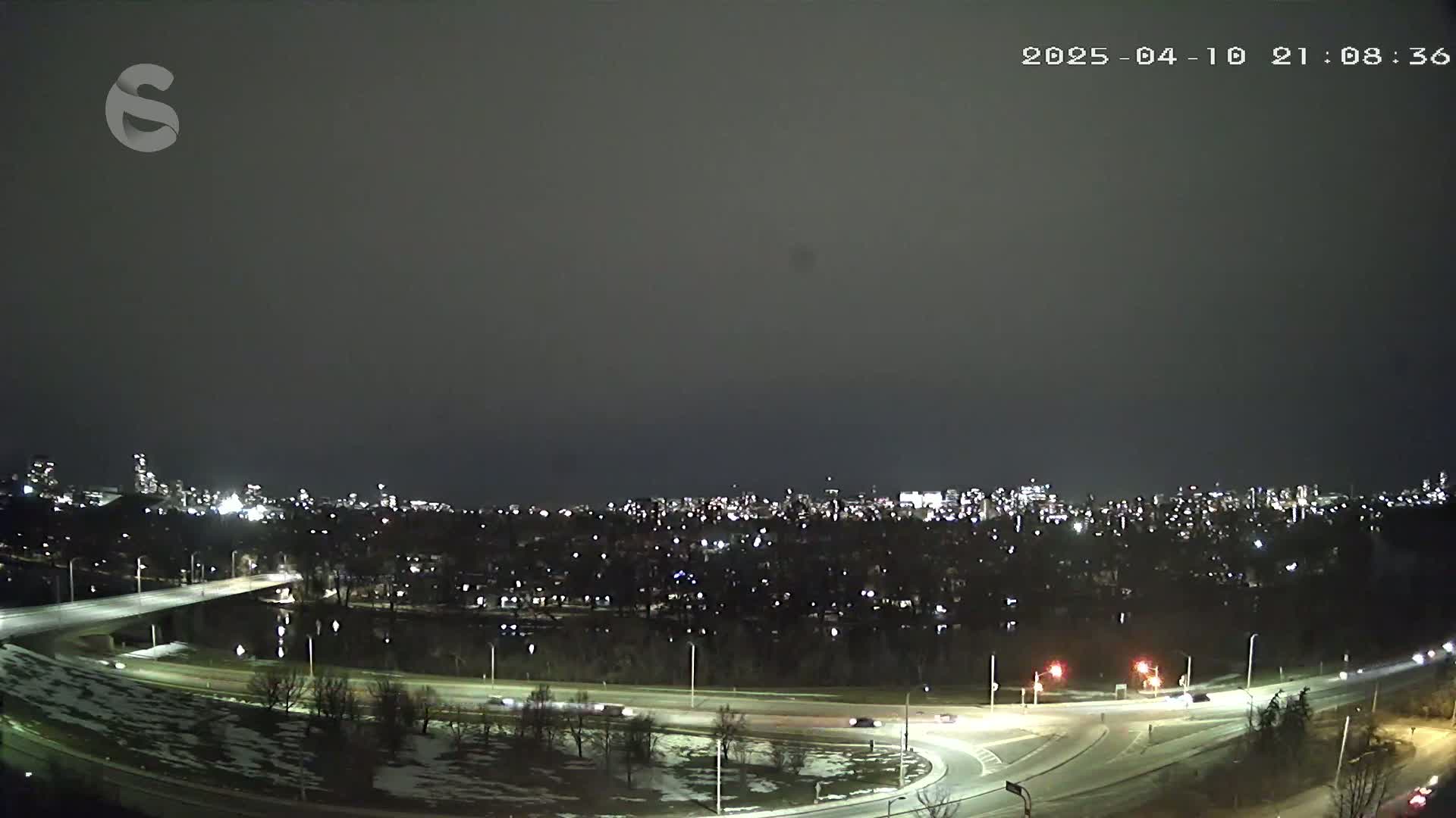 A nighttime view of a city skyline across a highway interchange, with patches of snow on the ground under a dark, overcast sky.