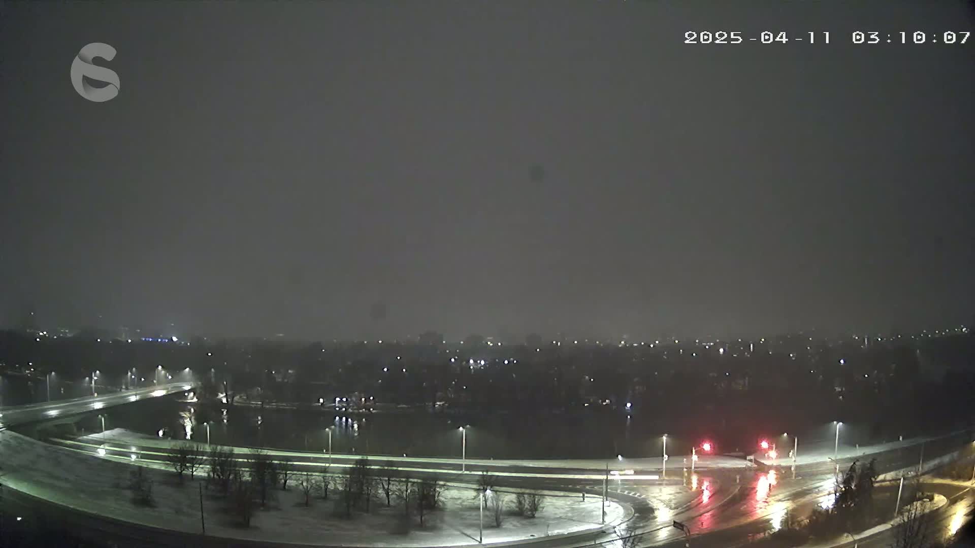 A nighttime, snowy scene shows a highway curving around a body of water, with dimly lit buildings and trees in the background.