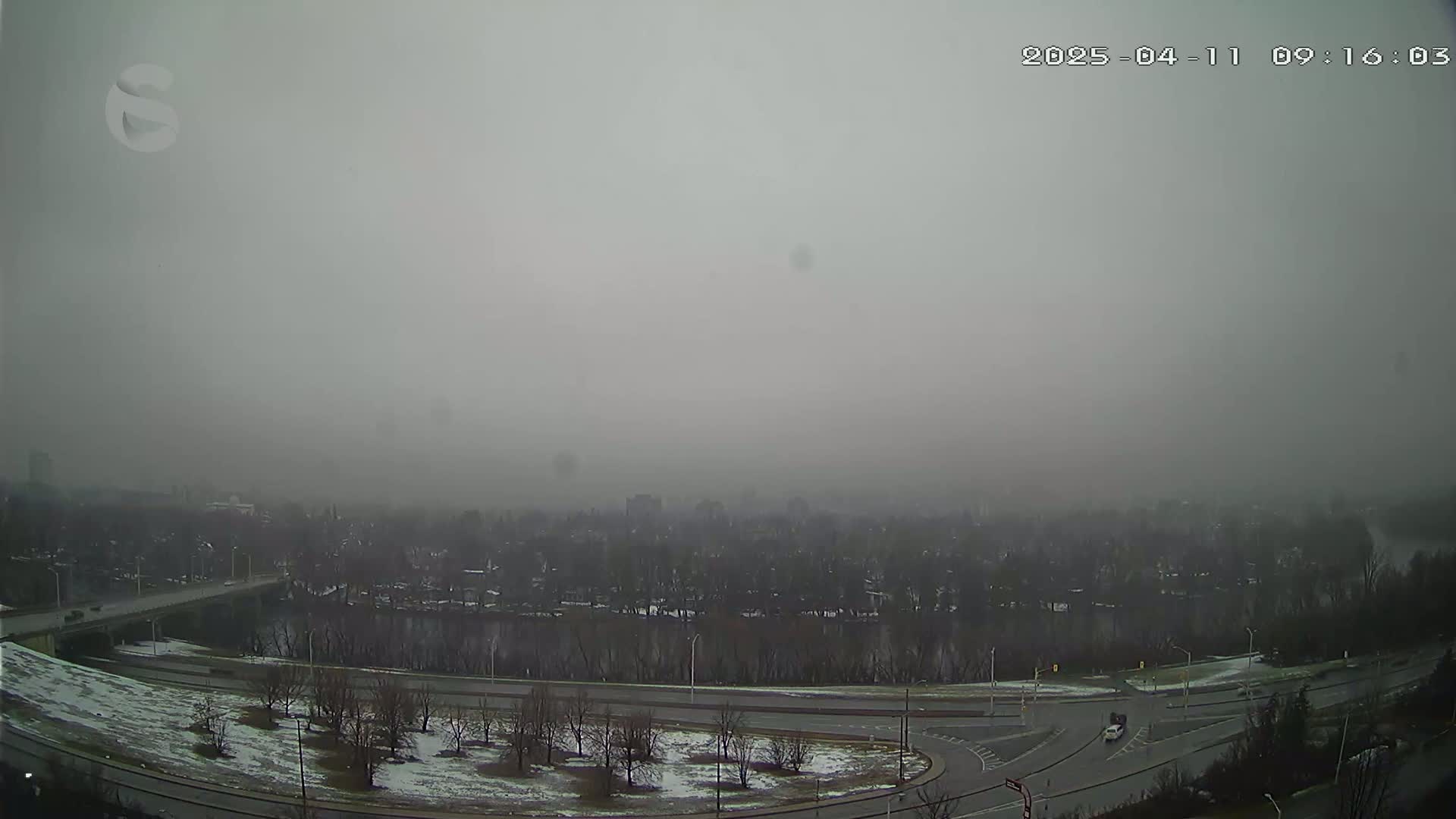 A foggy, overcast view shows a river winding through a residential area with a roadway and bridge, and patches of snow on the ground.