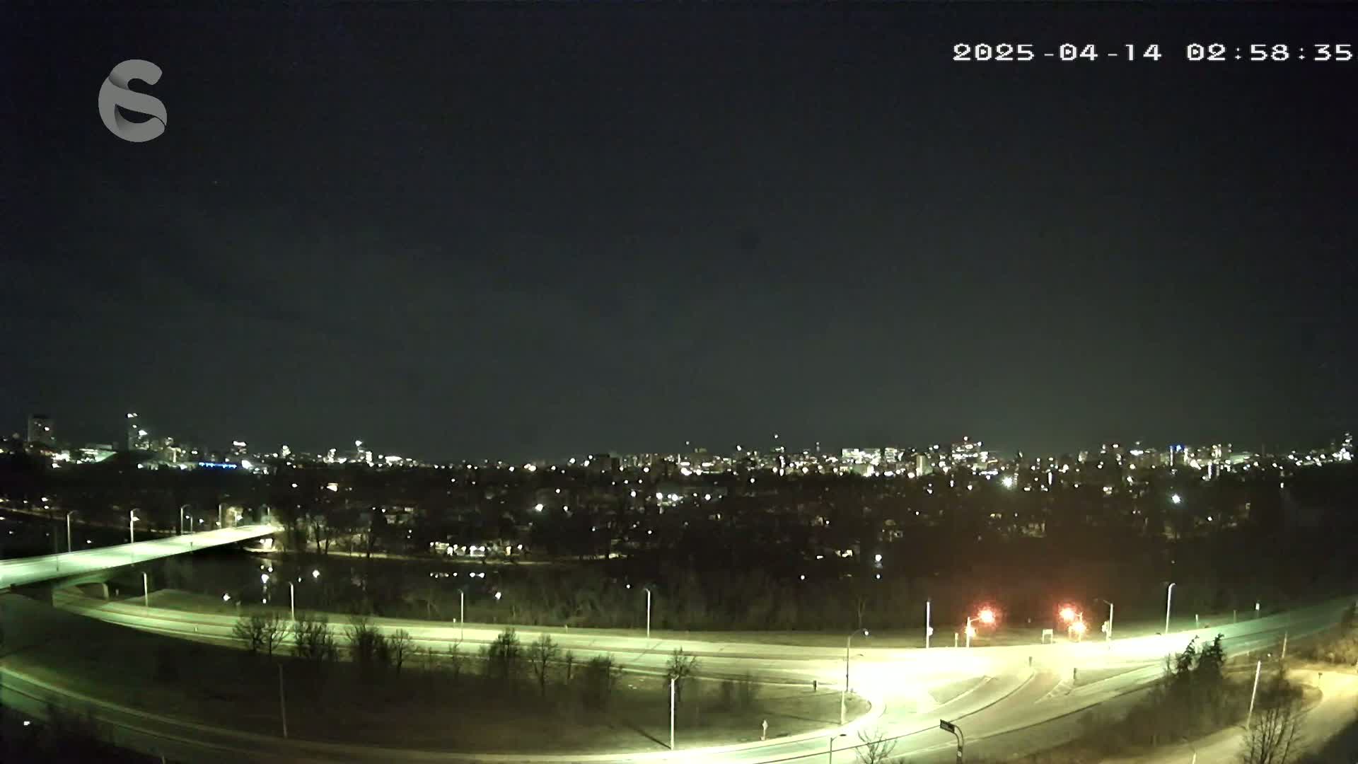 A nighttime view of a city skyline across a river, with illuminated roads and bridges under a clear, dark sky.