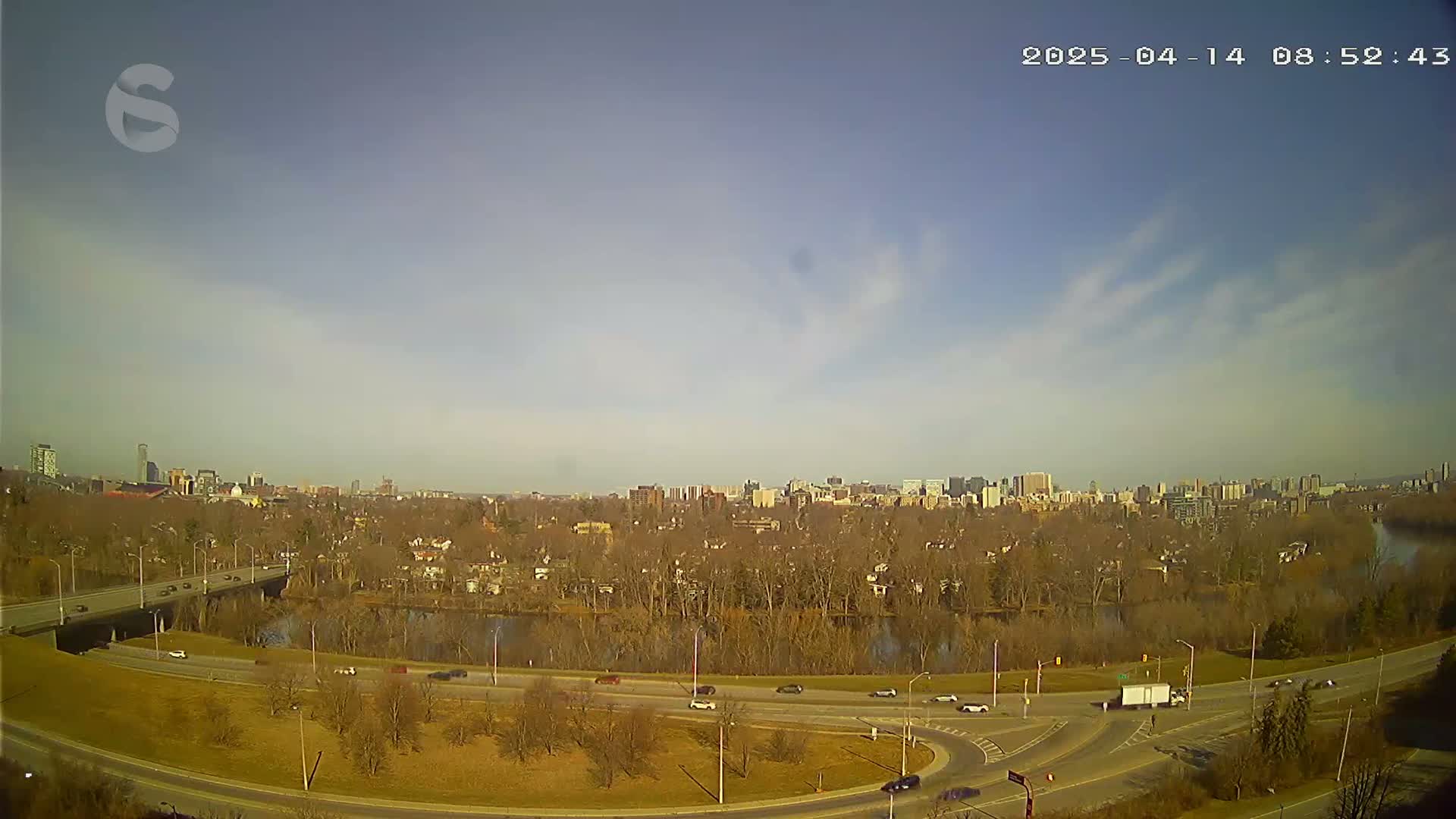 A sunny day reveals a highway curving around a river, with a city skyline visible in the distance and bare trees lining the riverbanks.