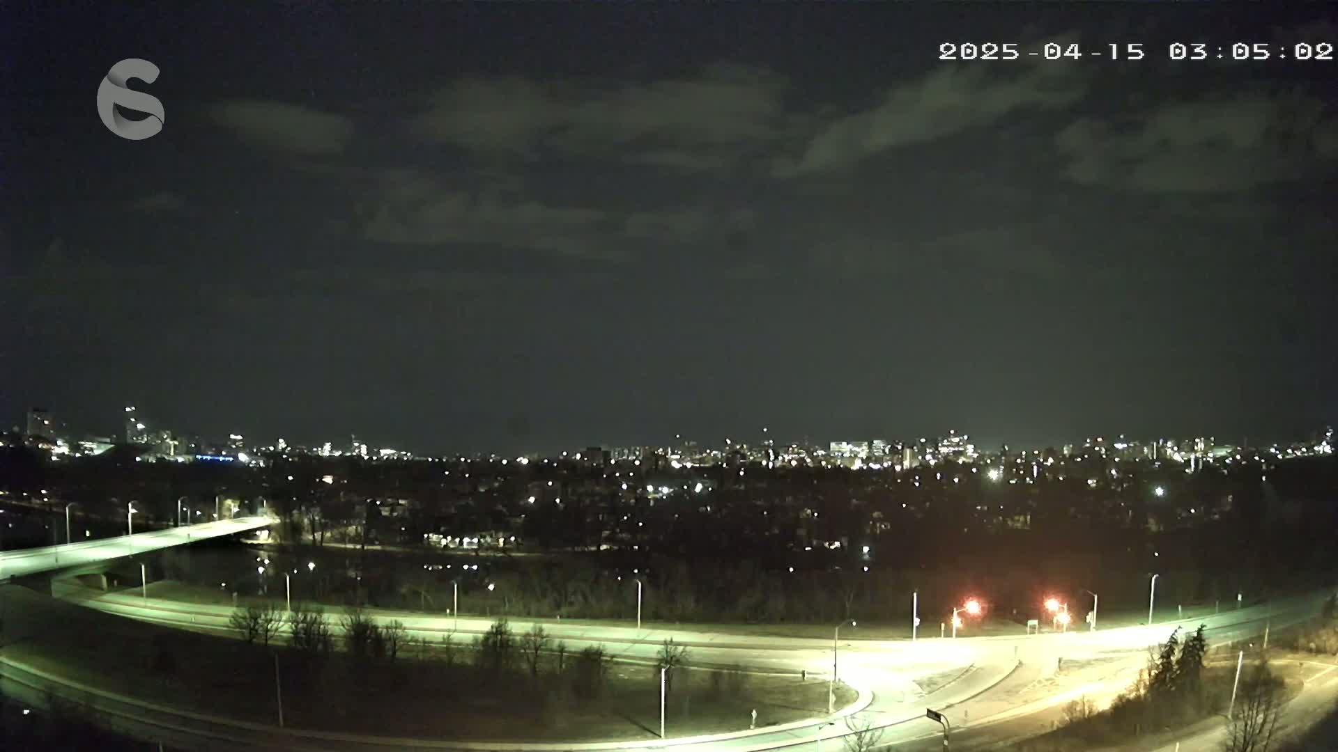 A nighttime view of a city skyline across a river, with a highway interchange in the foreground under a partly cloudy sky.