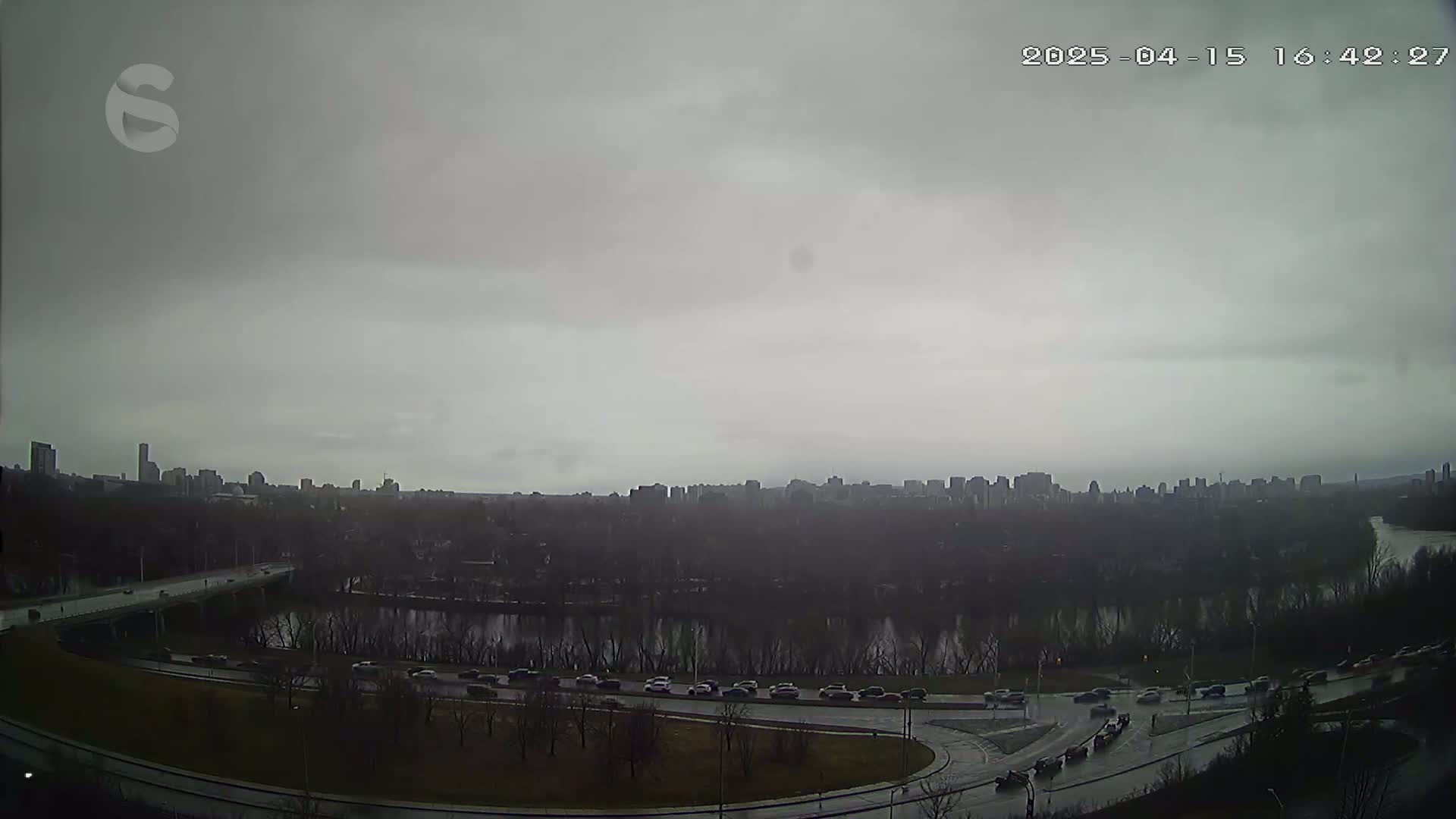 A city skyline is visible in the distance across a river under an overcast sky, with cars traveling on a highway and roads in the foreground.