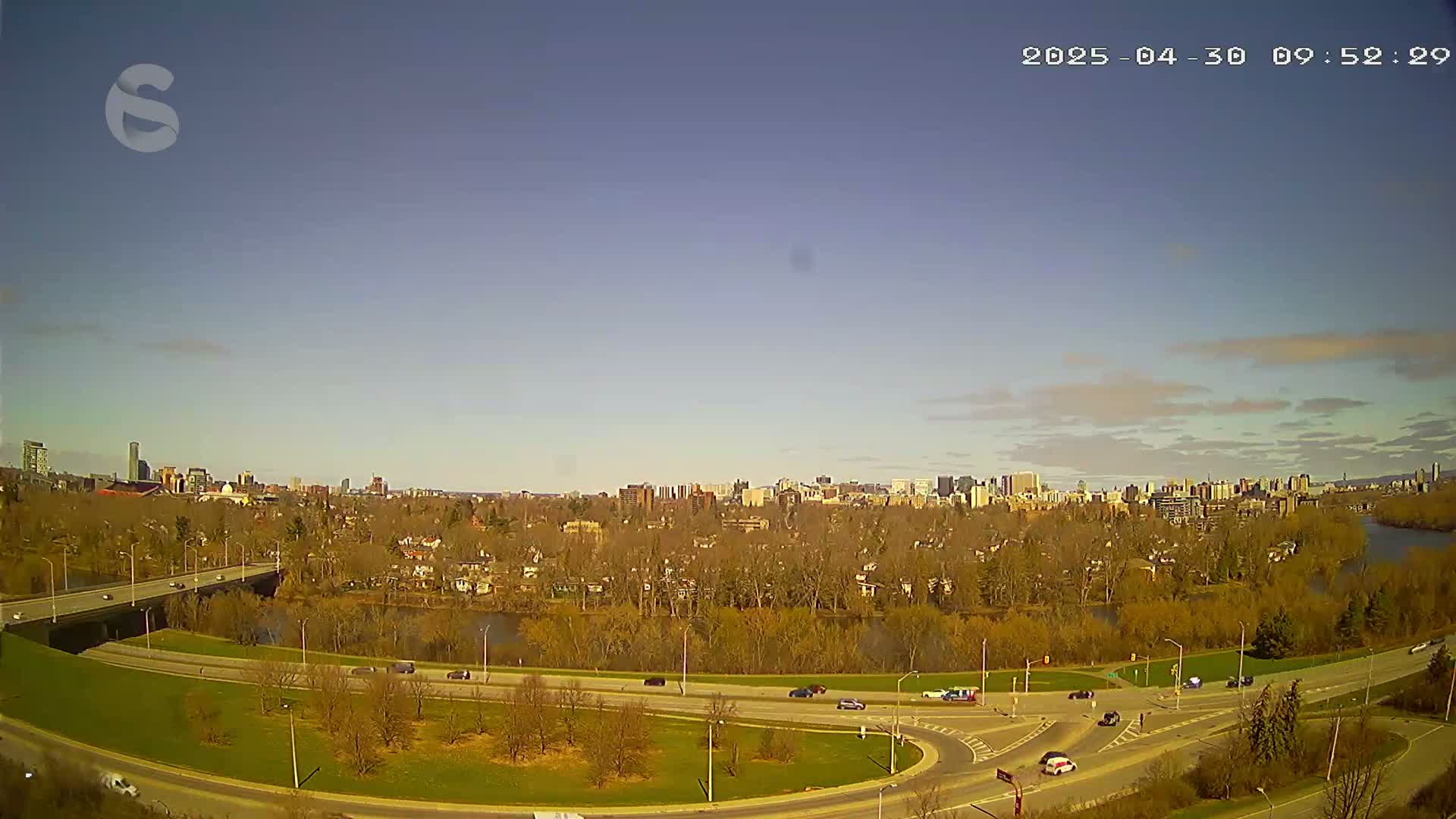 A clear, sunny day reveals a cityscape across a river, viewed from a high vantage point overlooking roads and bare trees.