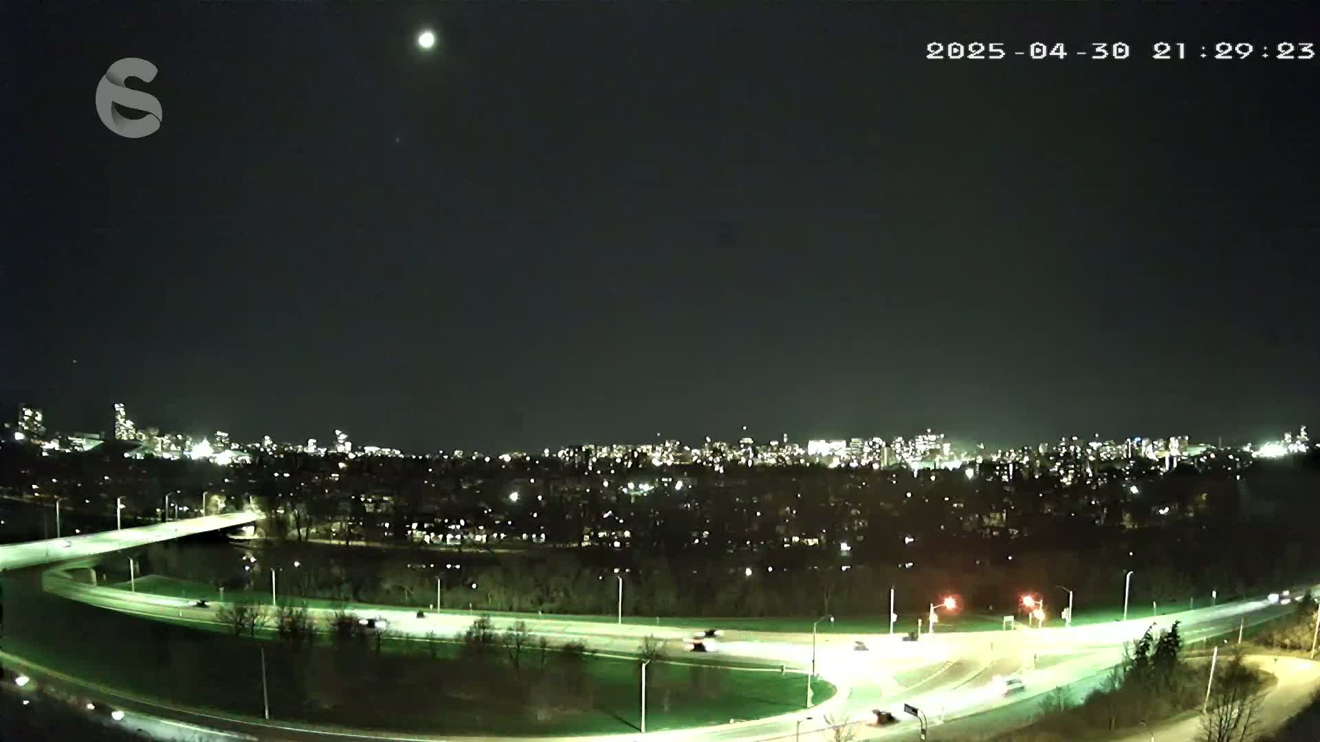 A nighttime, aerial view shows a highway interchange curving through a wooded area with a city skyline in the background under a clear sky.