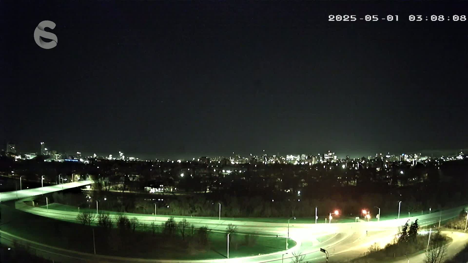 A nighttime, overhead view shows a highway interchange curving through a park-like area with a city skyline in the distance under a clear, dark sky.