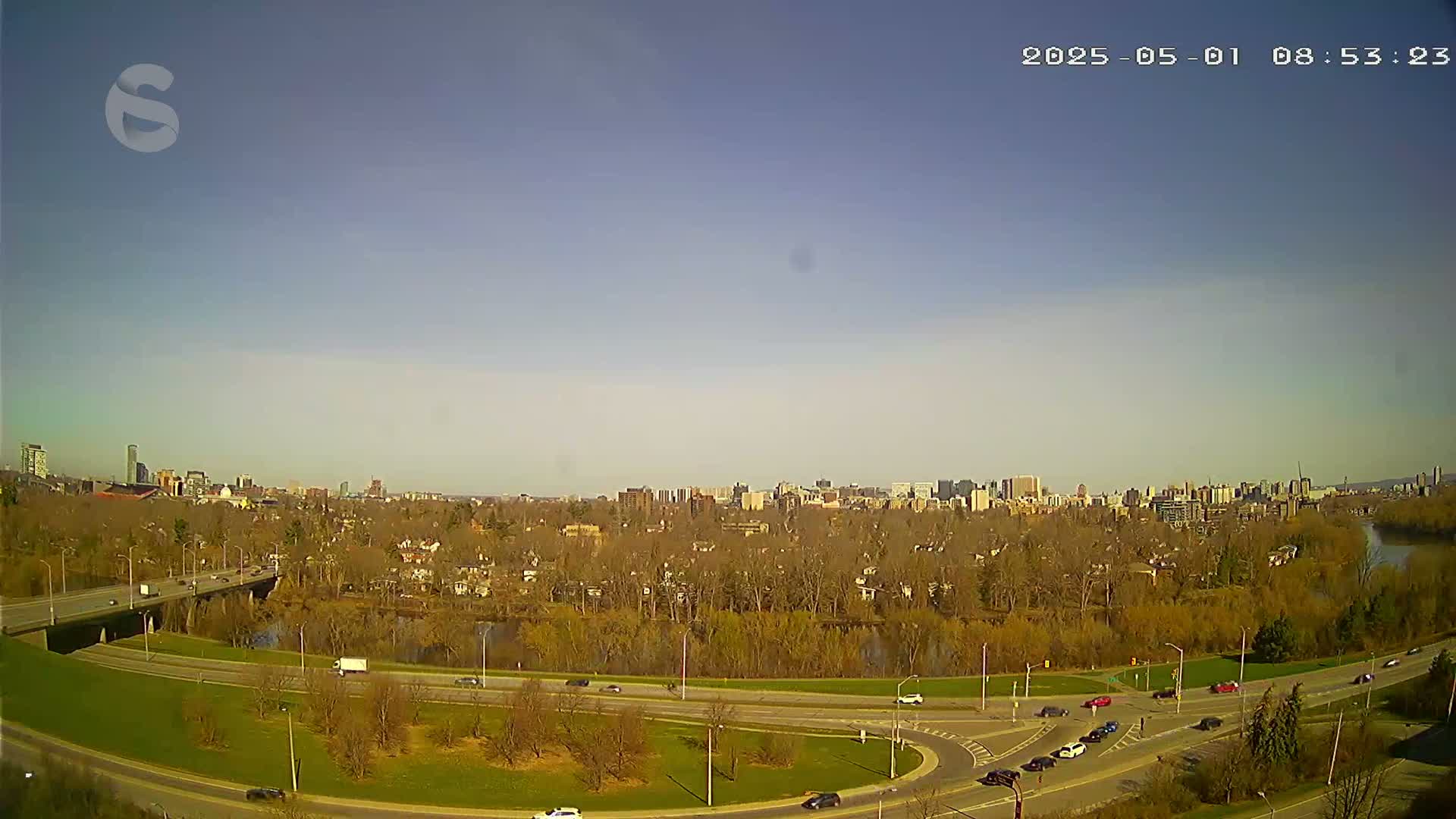 A clear, sunny day reveals a highway curving through a park-like area, with a city skyline visible in the distance along a river.