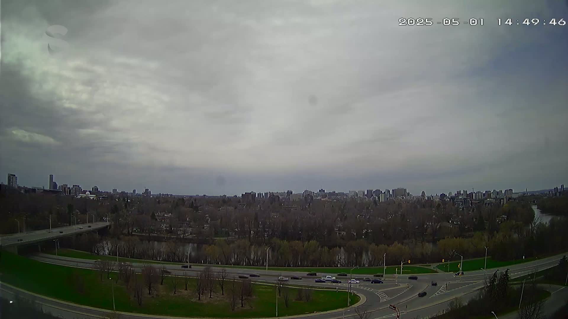 A city skyline is visible in the distance under an overcast sky, with a river and highway curving through a tree-lined landscape in the foreground.