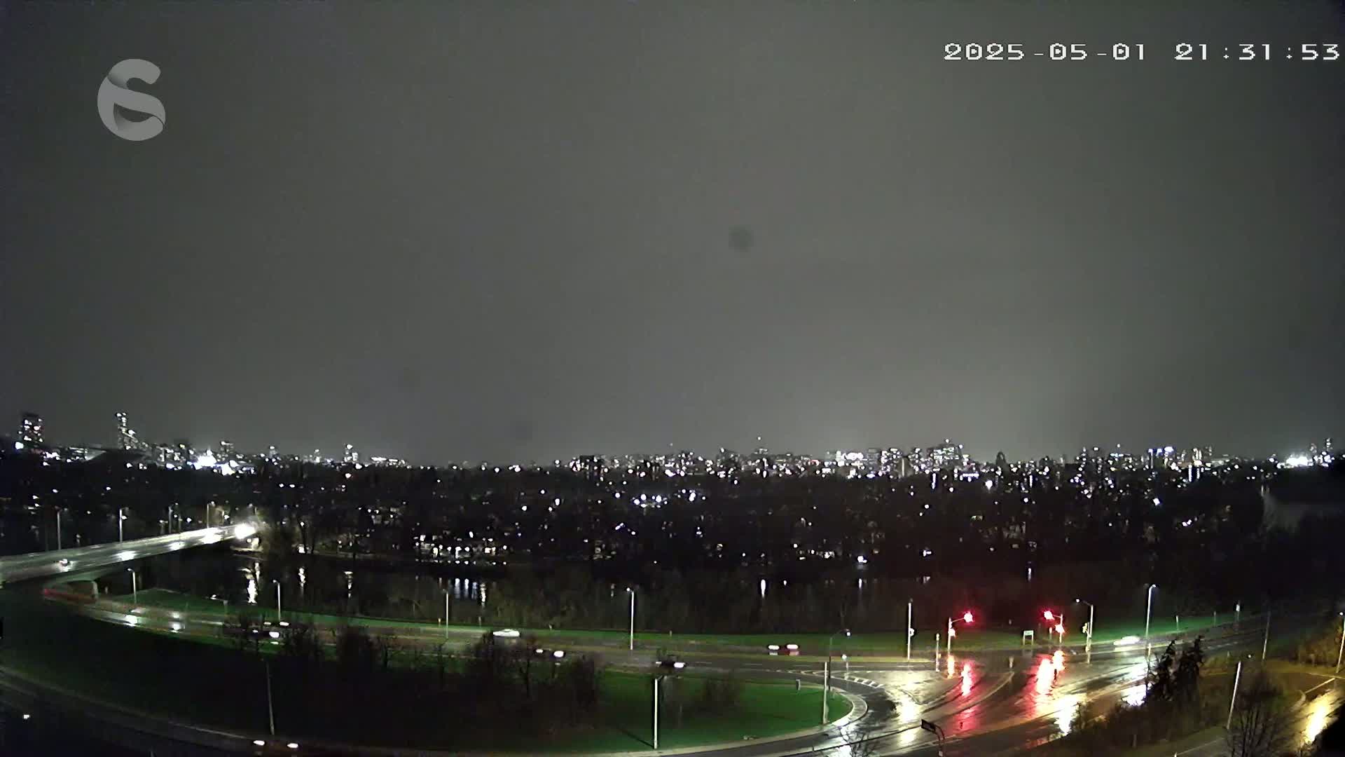 A nighttime view of a city skyline across a rain-slicked highway and roundabout.