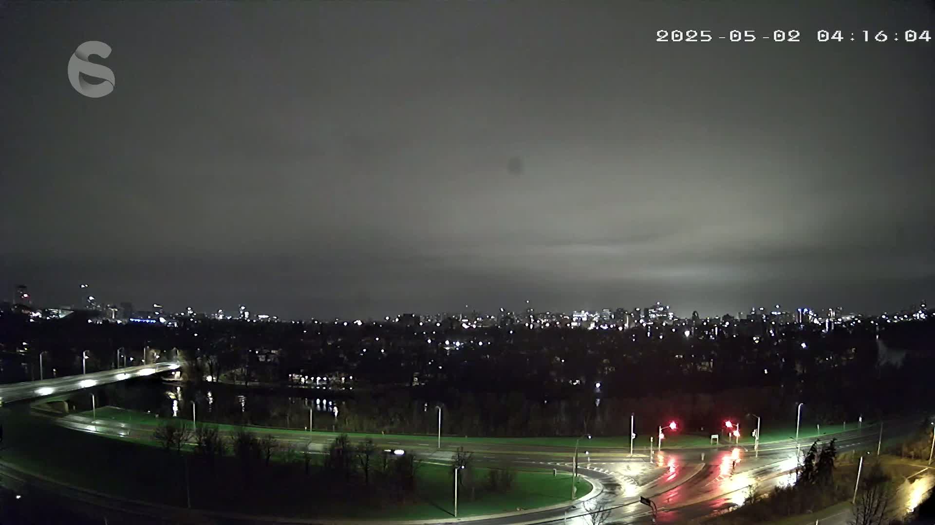 A nighttime view of a city skyline across a river, with wet roads and overcast skies.