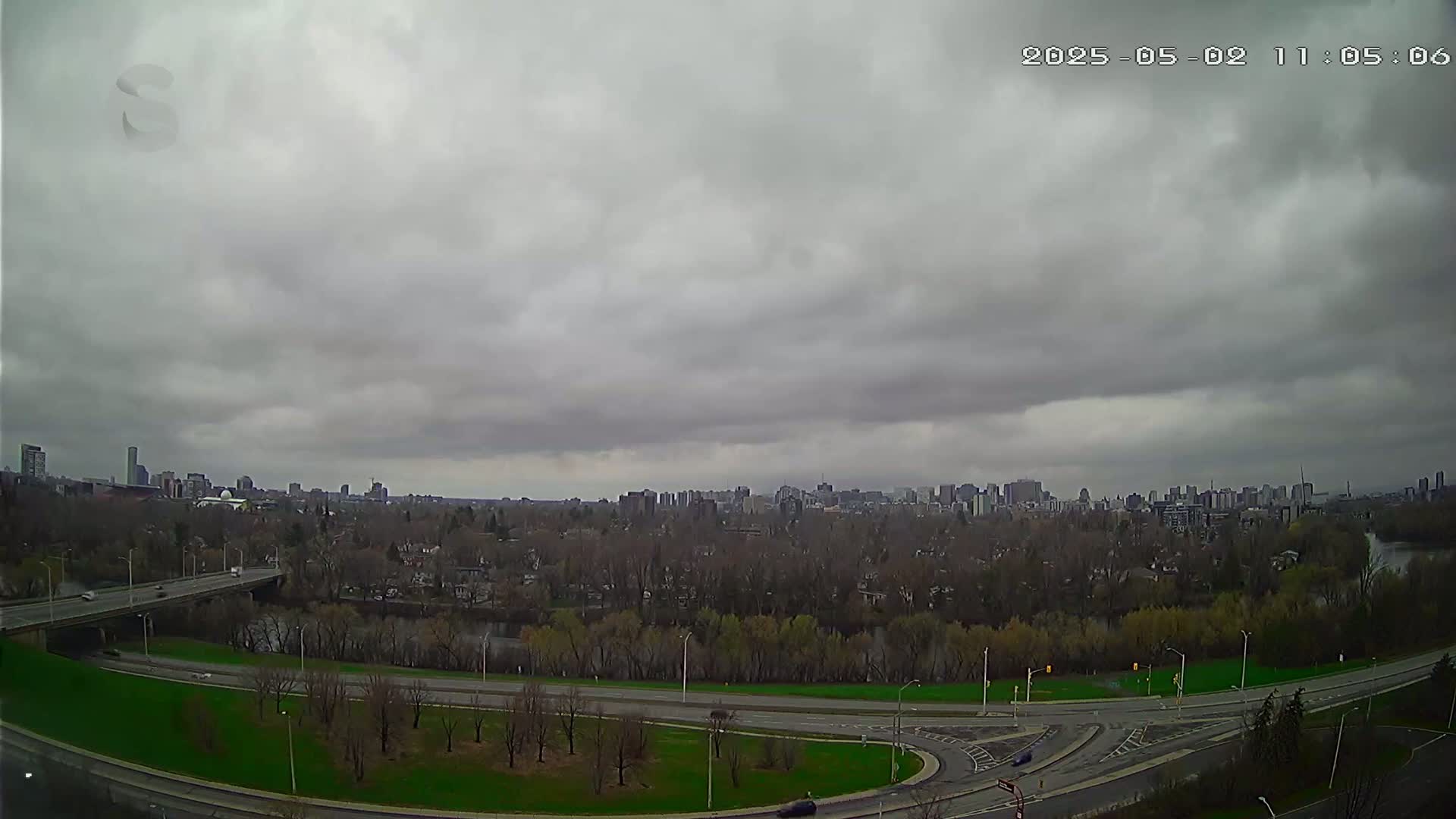 A city skyline is visible in the distance under a heavily overcast sky, with a highway curving through a park-like area in the foreground.