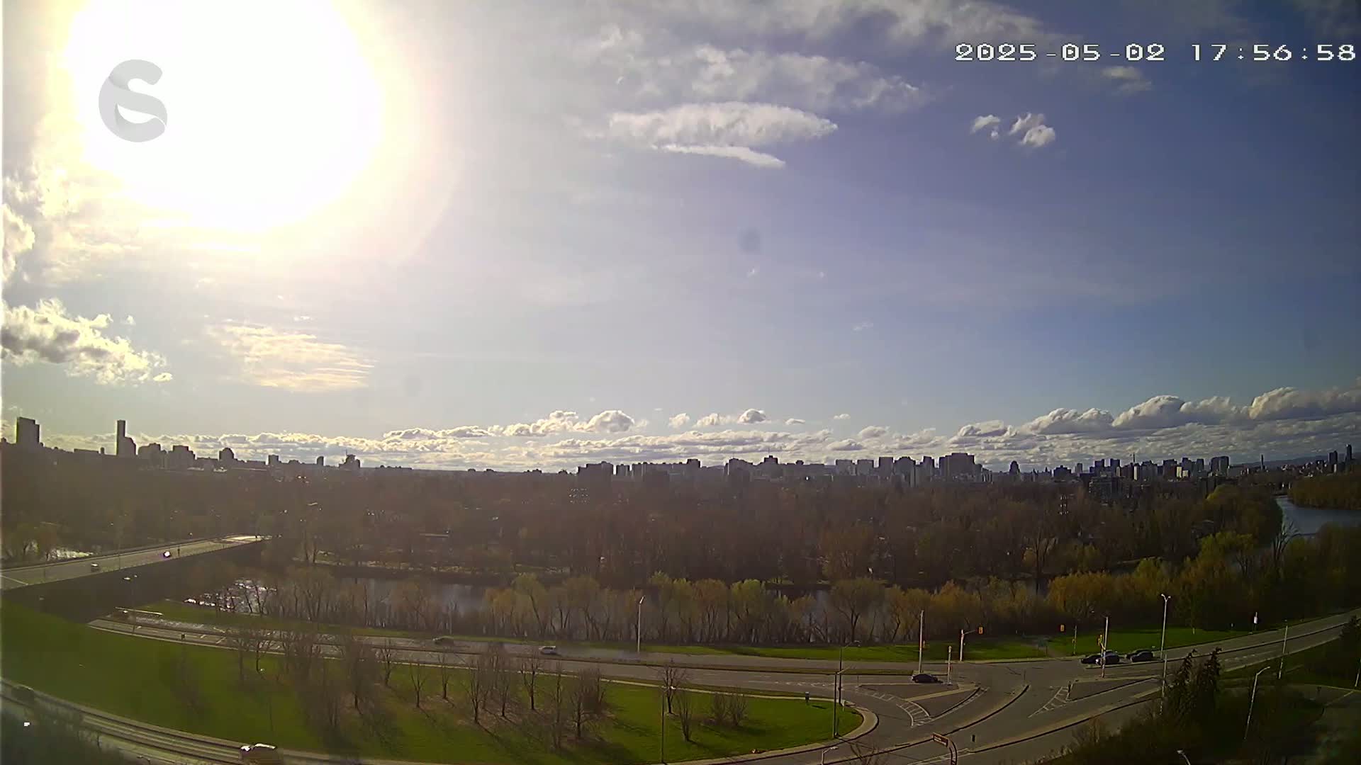 A sunny day with partly cloudy skies reveals a city skyline in the distance, viewed from an elevated vantage point overlooking a river, a bridge, and a roadway with sparse traffic.