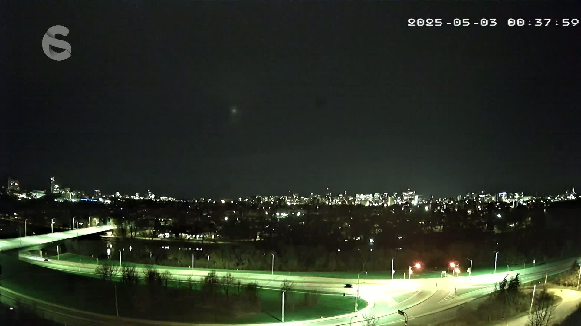 A nighttime, high-angle view shows a highway interchange curving through a dark, tree-lined area toward a city skyline illuminated by lights.