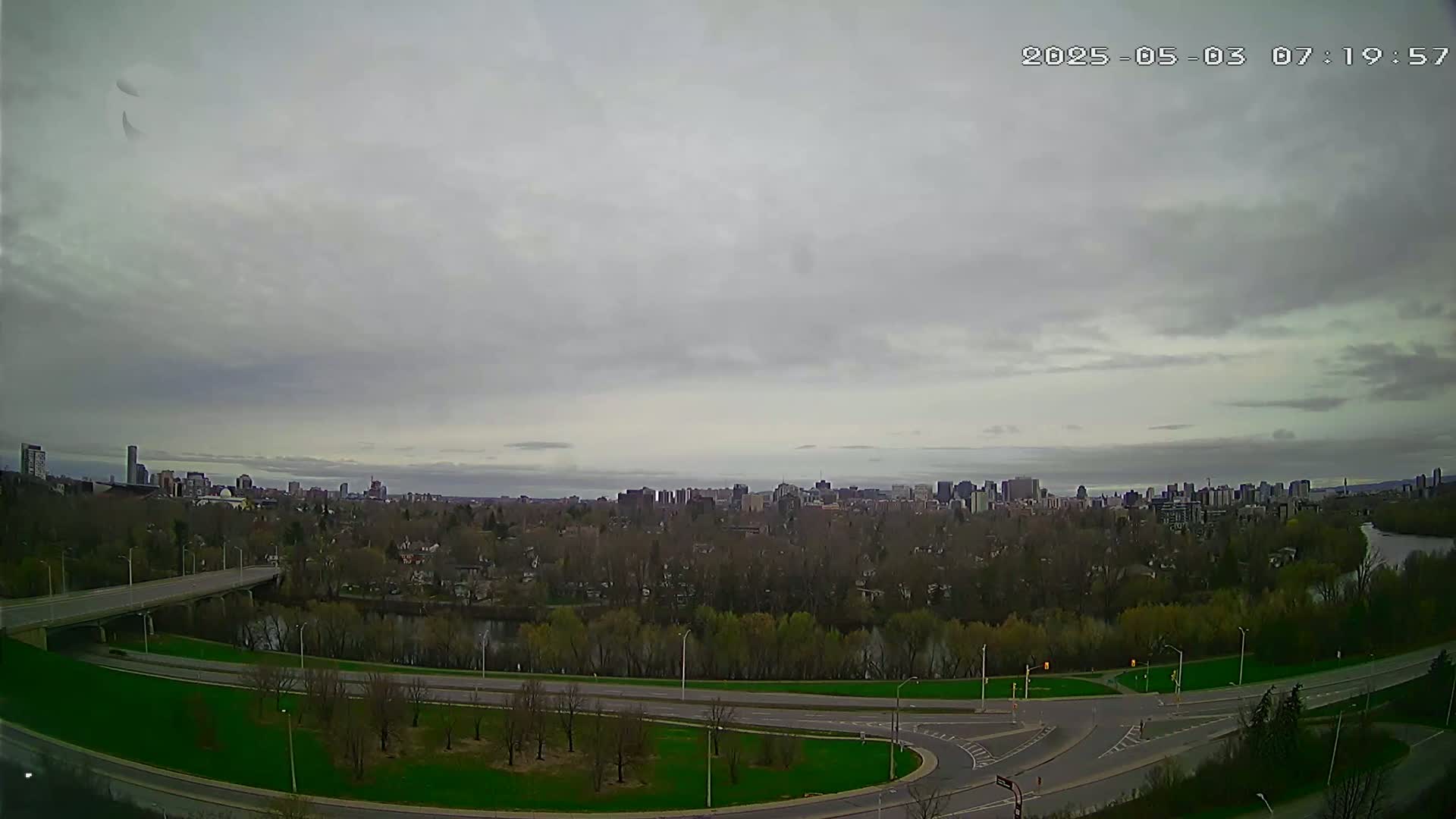 A wide shot of a city skyline on an overcast day, viewed from across a river and highway system with green spaces in between.