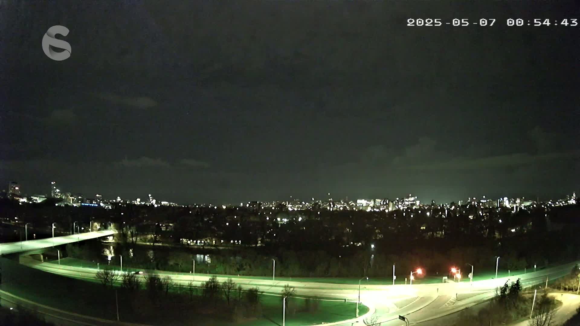 A nighttime view of a city skyline across a river, with a highway curving through the foreground under a partly cloudy sky.