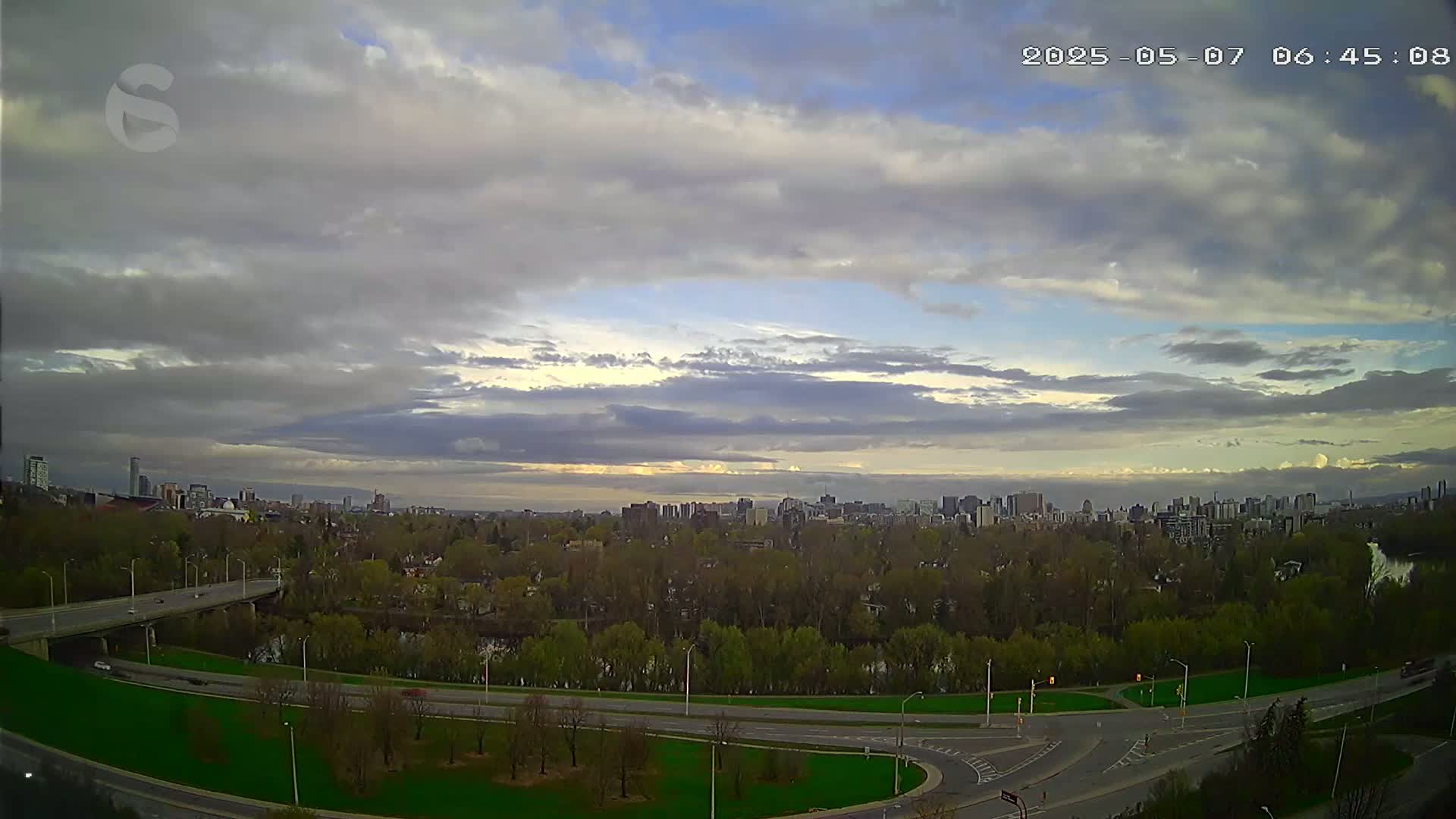 A partly cloudy sky over a city skyline and a river bordered by trees and roads.