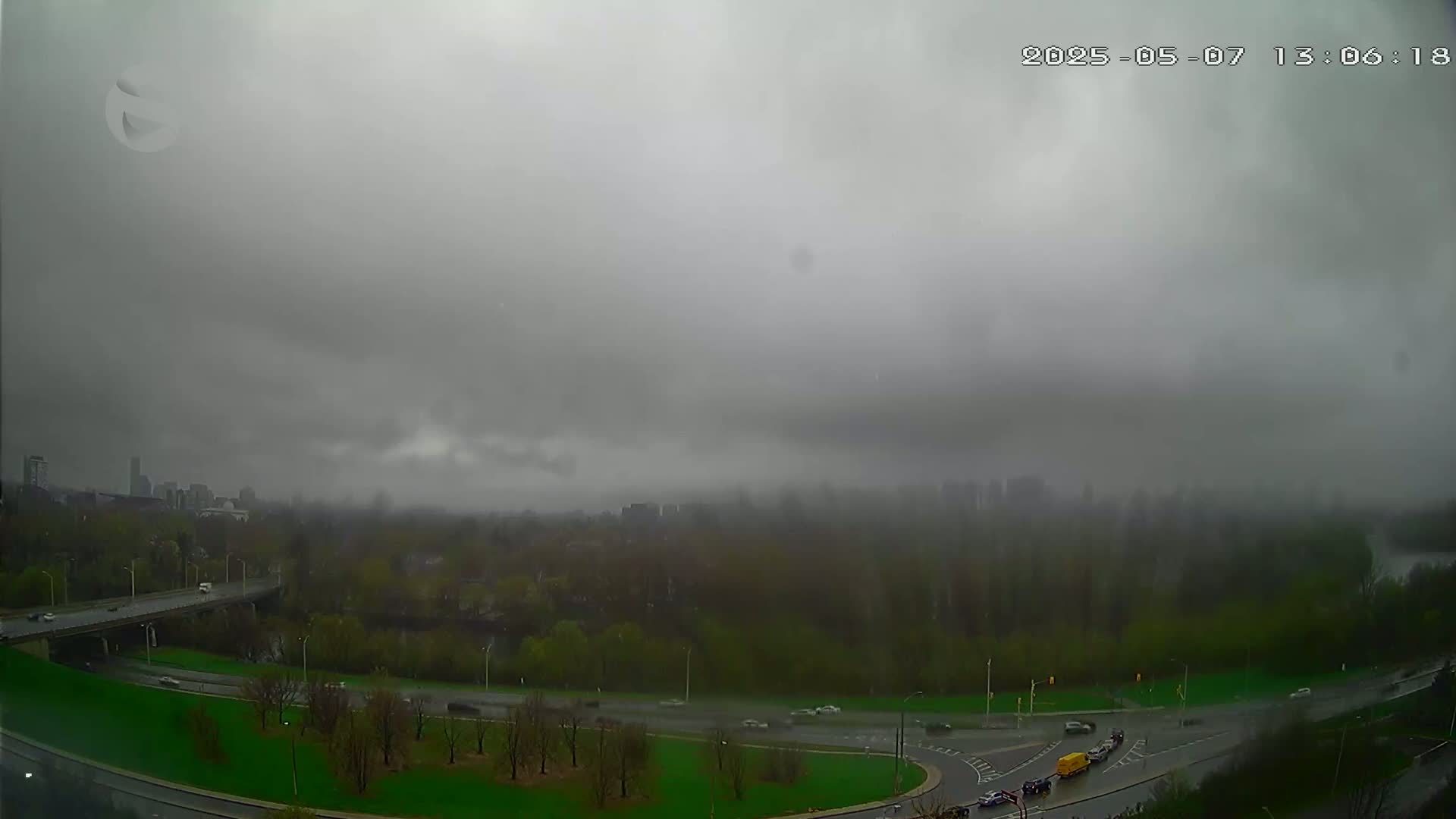 A hazy, overcast day reveals a highway system curving through a green space beside a river, with buildings and trees visible in the distance under a thick layer of clouds.