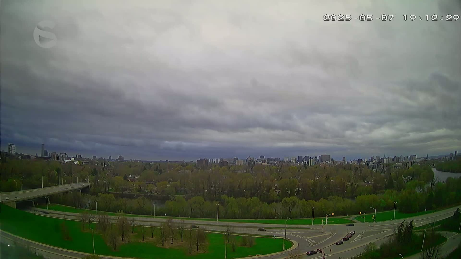 A sprawling cityscape is visible in the distance under a cloudy sky, with a highway curving through a green, tree-lined area in the foreground.