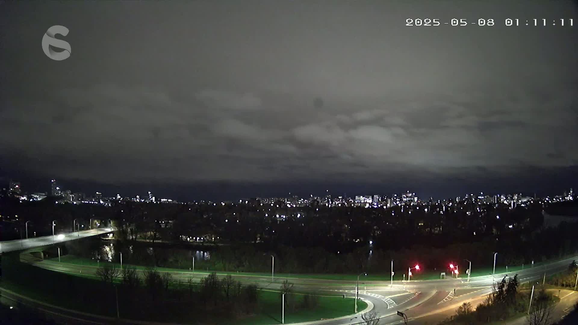 A nighttime view of a city skyline across a river, under a cloudy sky, shows a road curving through a park-like area near a bridge.