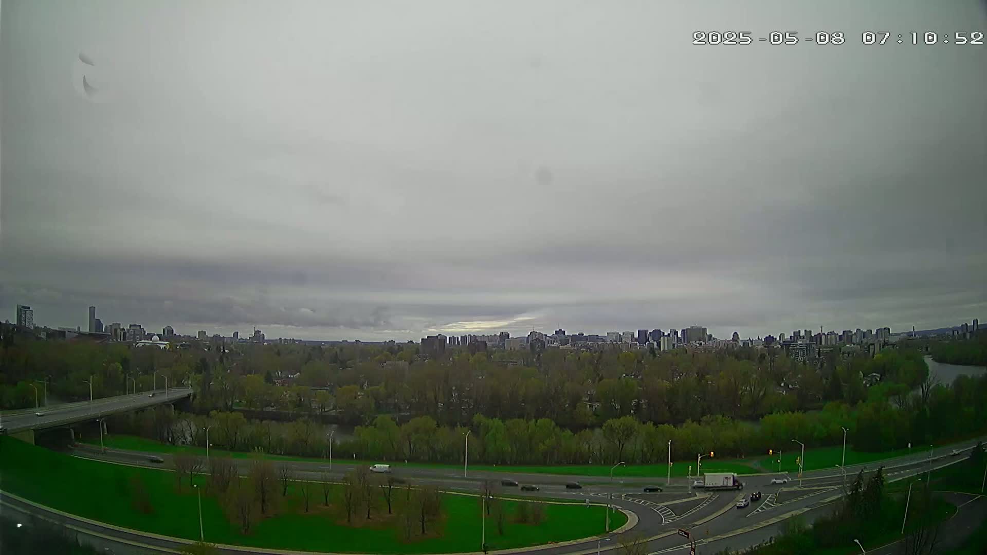 A high-angle view shows a cityscape on a cloudy day, with a river and highway visible in the foreground.