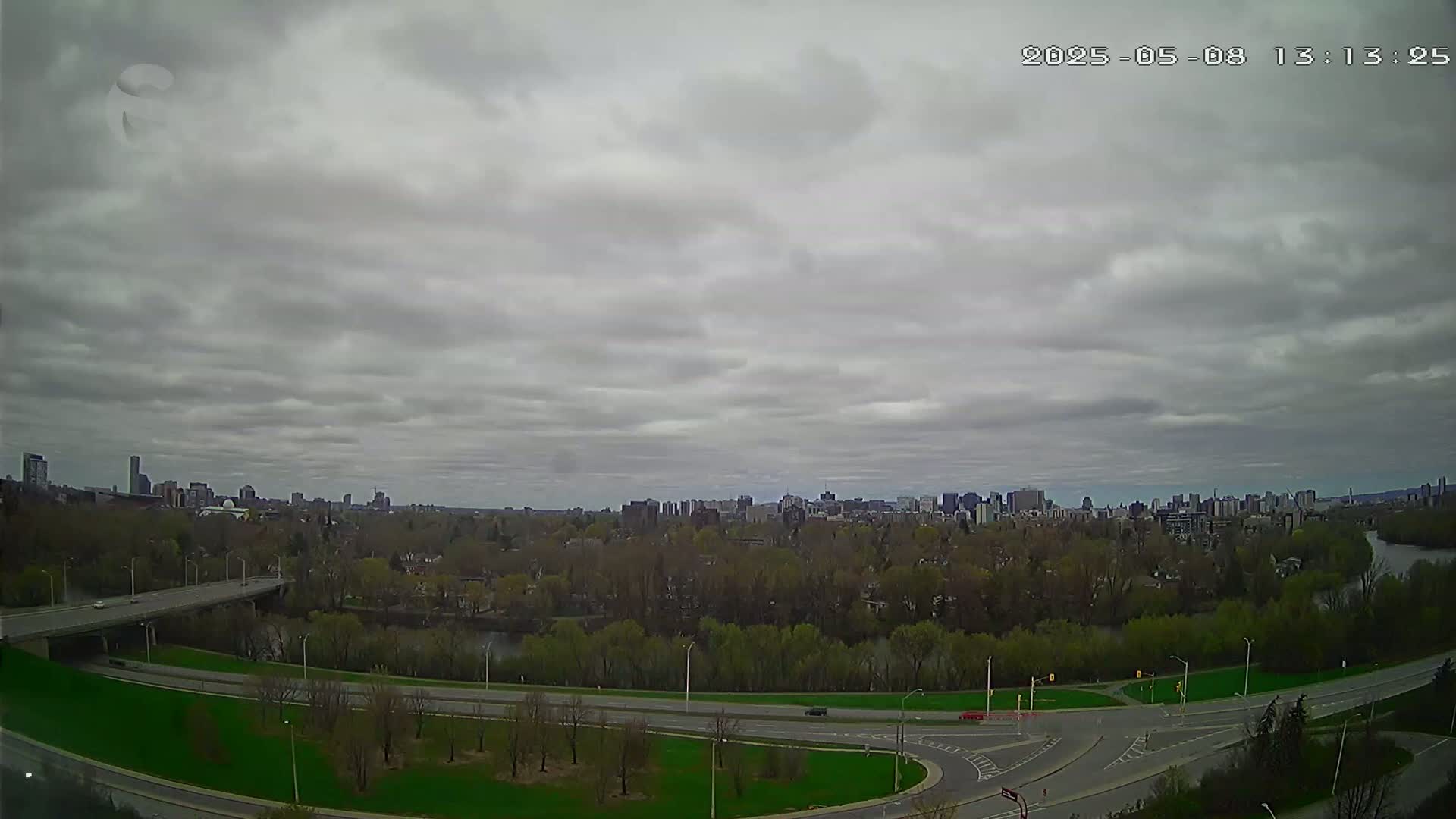 A cityscape under a cloudy sky is viewed from above, showing a highway system intersecting near a river that runs through a green, tree-lined area.