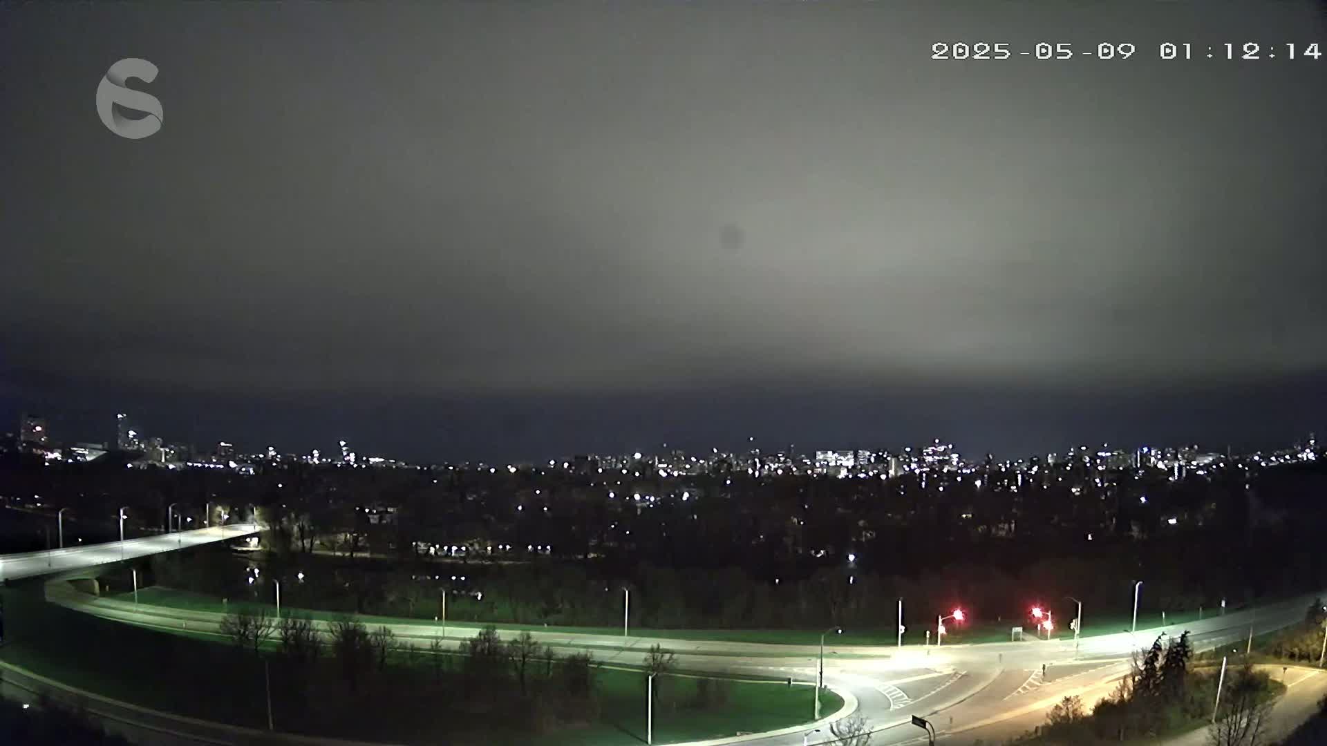 An elevated nighttime view of a city skyline across a dark, overcast sky, with a curved roadway and bridge in the foreground.