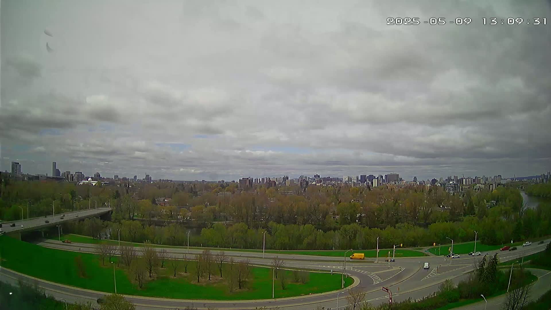 An elevated view shows a highway interchange curving through a green space with a city skyline in the background under a cloudy sky.
