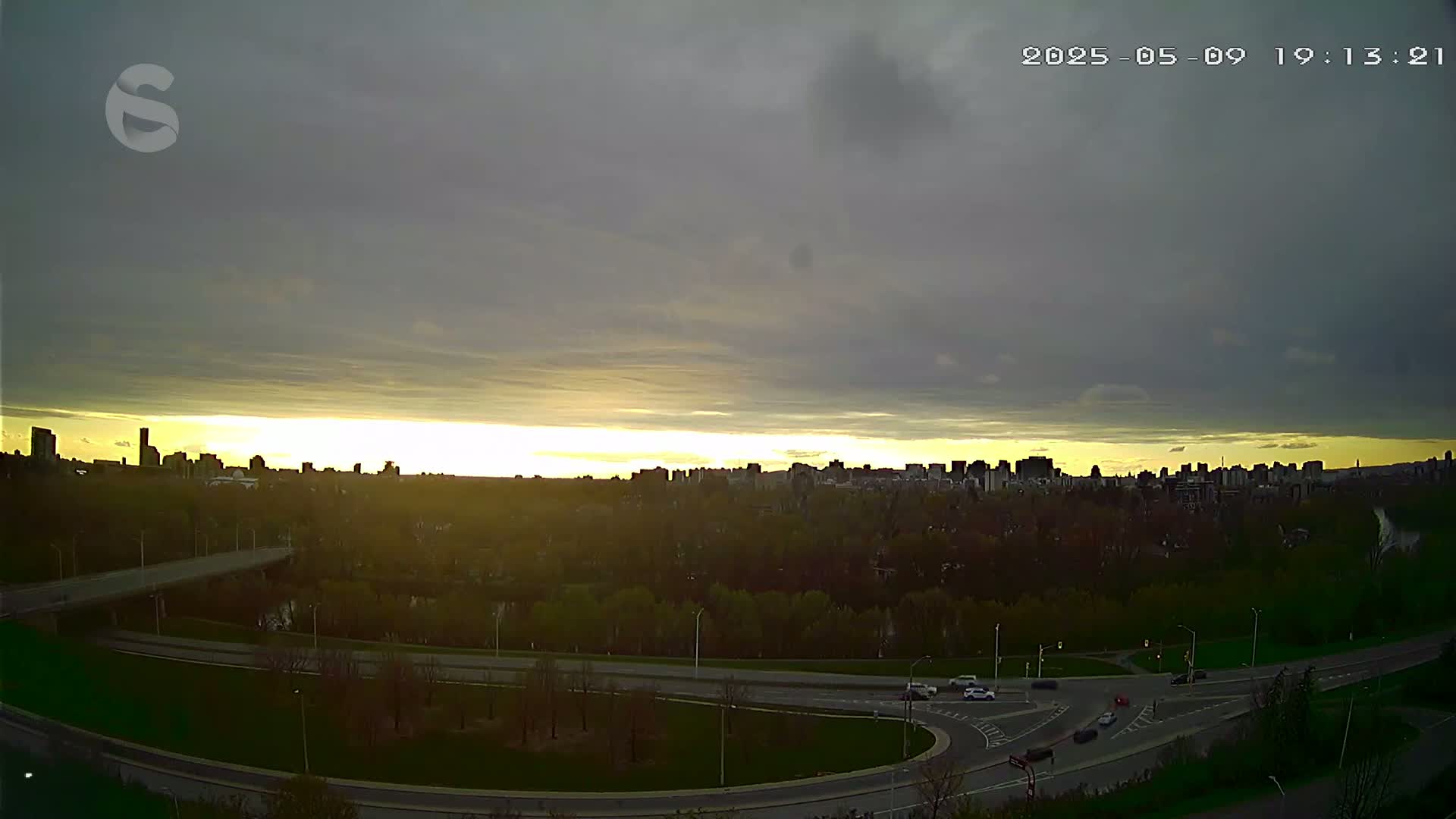 A city skyline is visible at sunset under a mostly cloudy sky, with a highway and green space in the foreground.