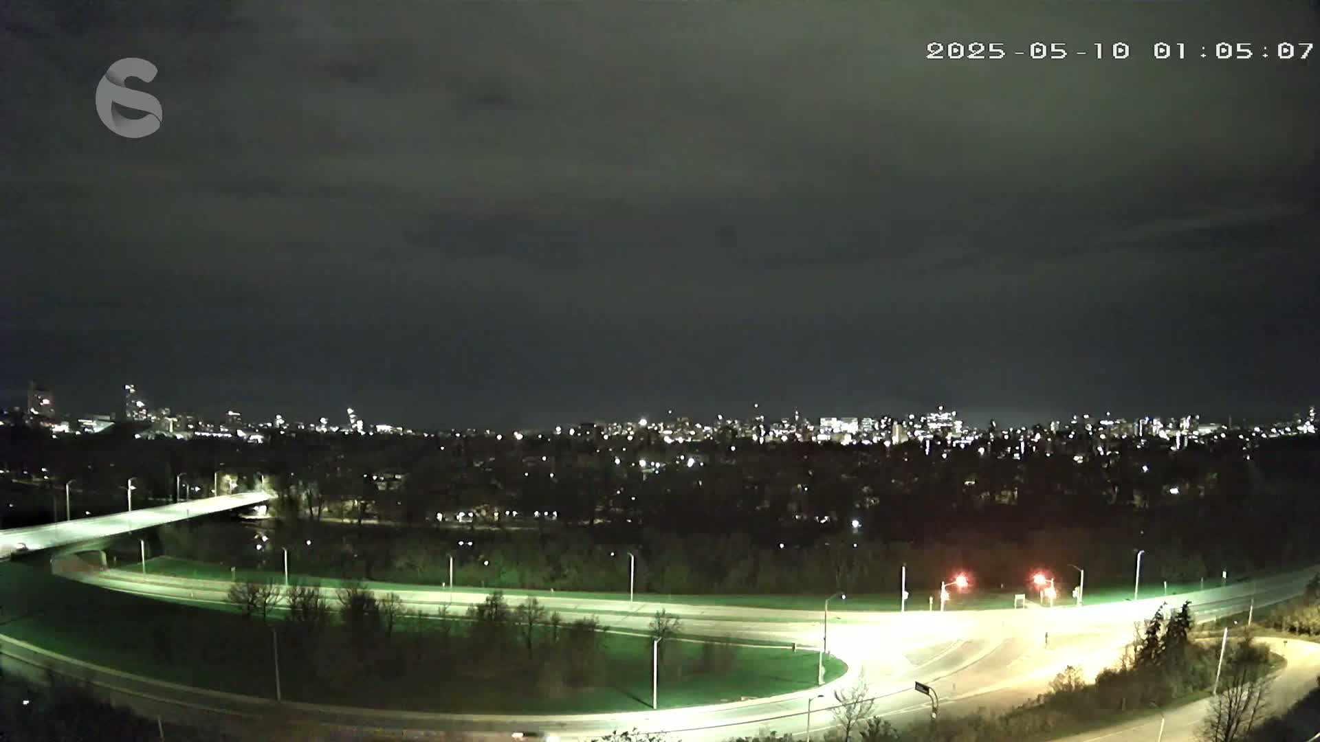 A nighttime view of a highway interchange and a city skyline under a dark, cloudy sky.