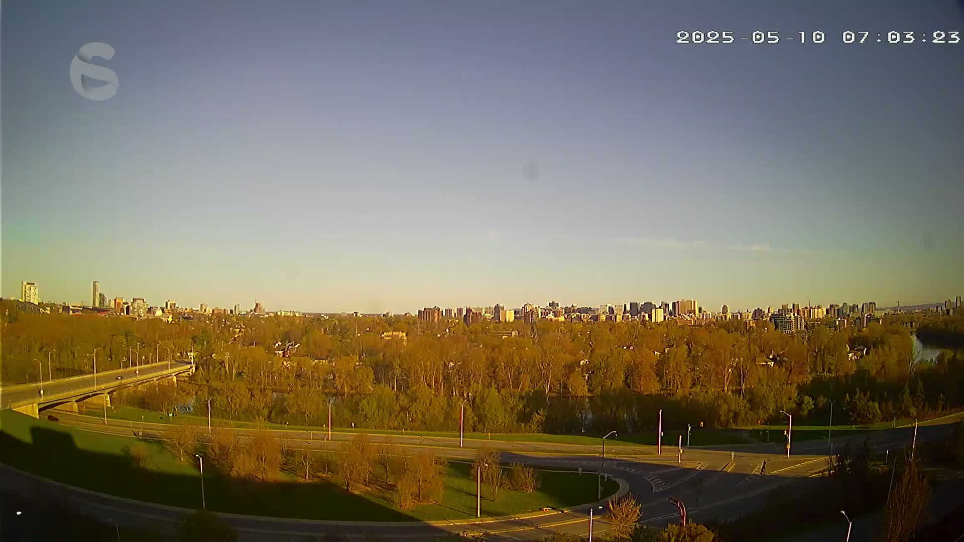 A cityscape is visible in the distance, beyond a highway and a tree-lined riverbank, under a clear, pale blue sky.