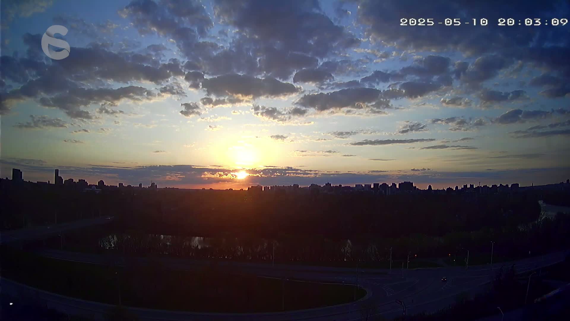 A partly cloudy sunset illuminates a city skyline beyond a river and highway.