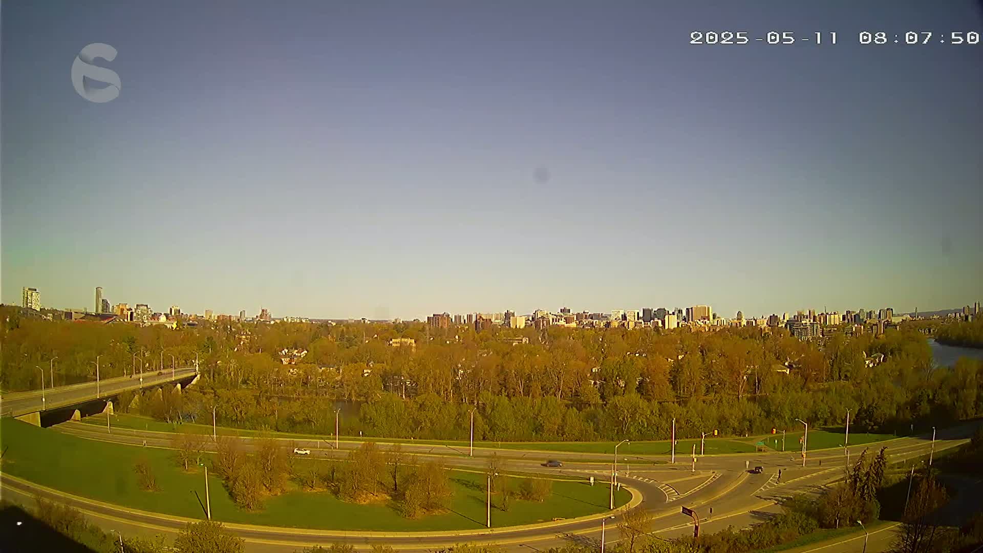 A clear, sunny day reveals a cityscape in the distance, viewed from above a highway interchange surrounded by a park-like setting with budding trees.