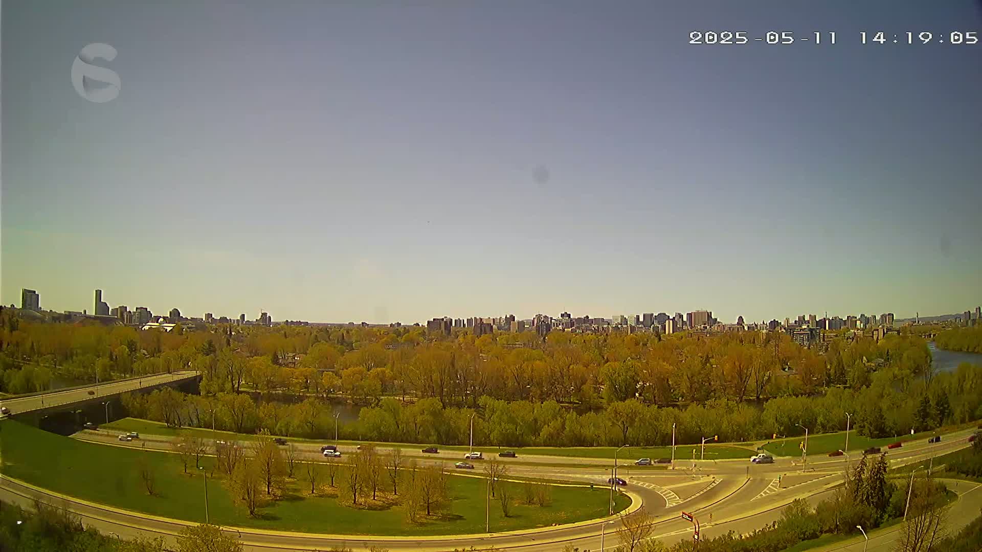 A cityscape is visible in the distance across a river, under a clear sky, with a roadway and green space in the foreground.