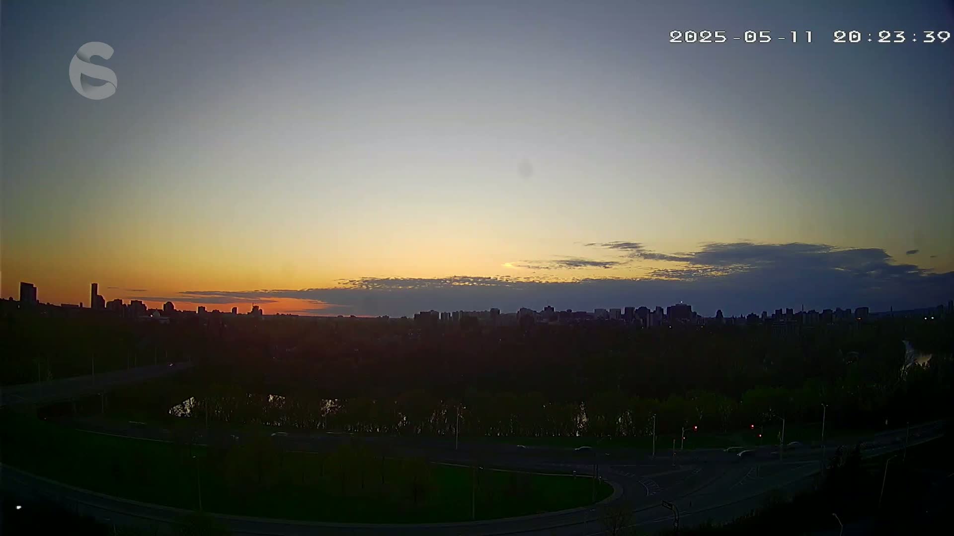 A city skyline silhouetted against a sunset sky with a few clouds, viewed from across a roadway and a river.
