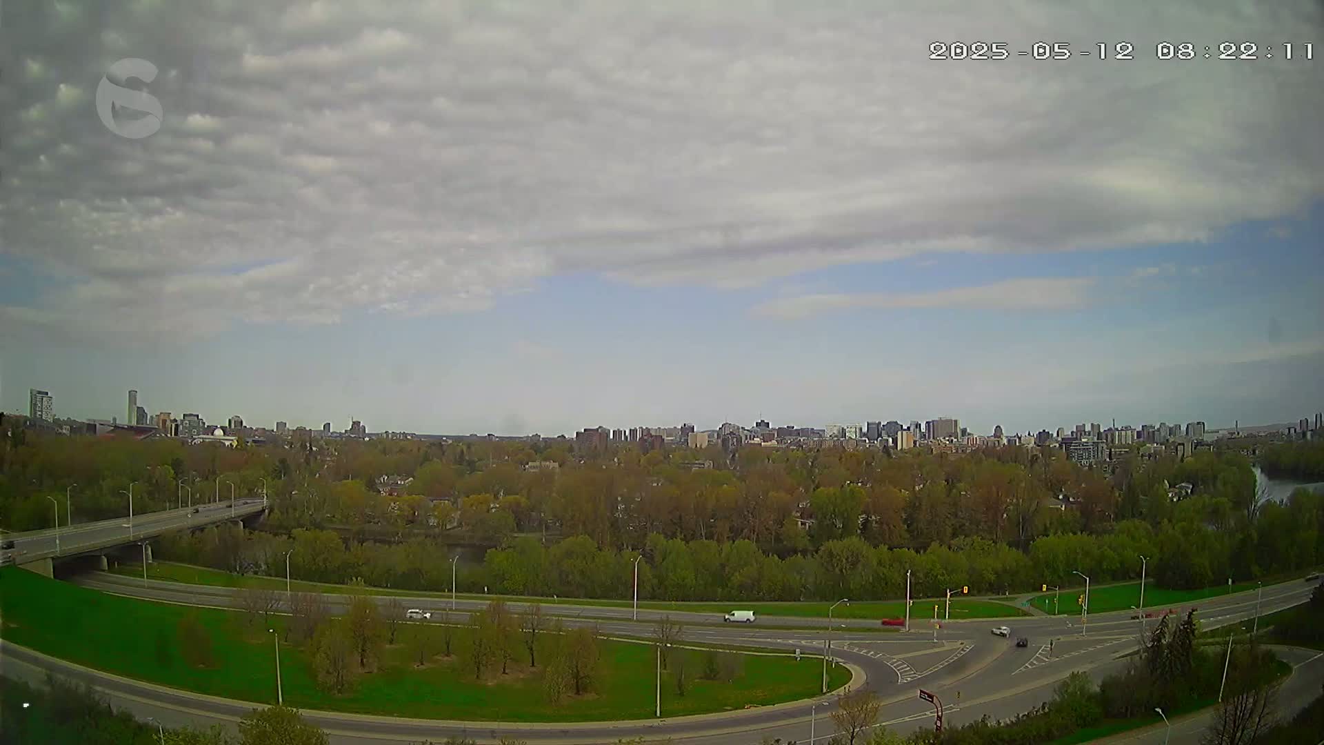 A city skyline is visible in the distance under a partly cloudy sky, with a highway and a river visible in the foreground.