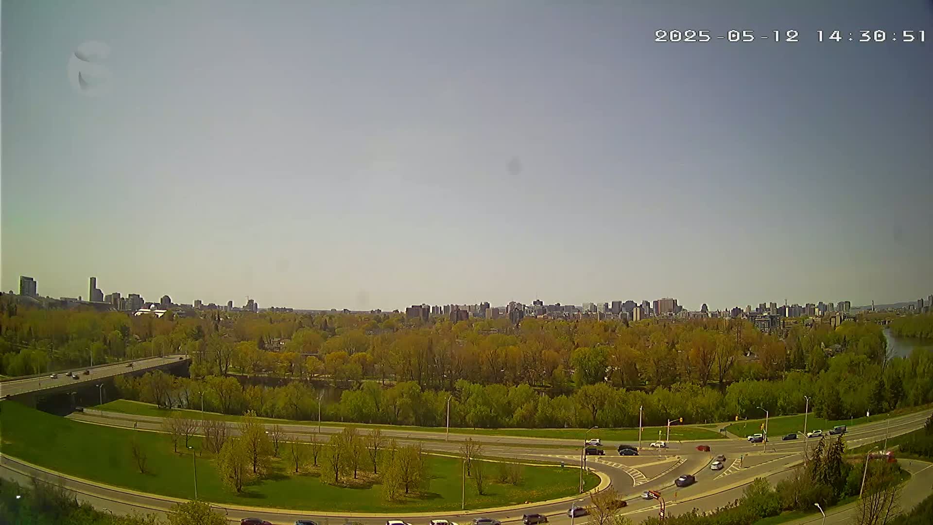 A mostly sunny cityscape is visible beyond a verdant expanse of trees and a roadway system, including a bridge.