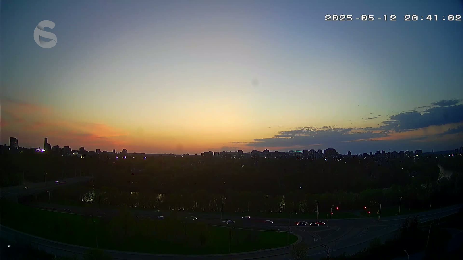 A city skyline at sunset is viewed from above a roadway, with a few cars visible, under a mostly clear sky.