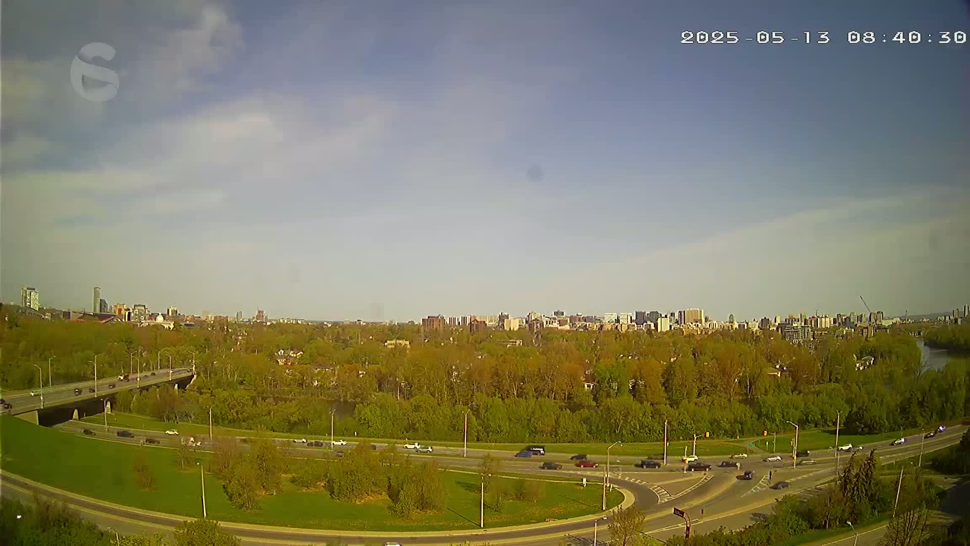 A partly cloudy sky looks down on a highway overpass and surrounding roads, green trees, and a distant cityscape.