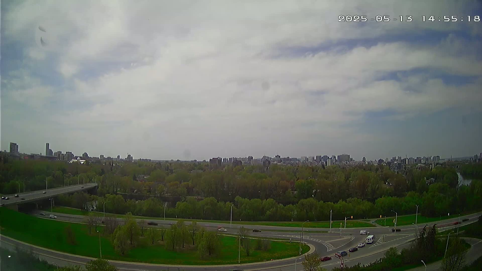 A partly cloudy sky overlooks a city skyline and a highway curving through a green, tree-lined area with several vehicles visible on the road.