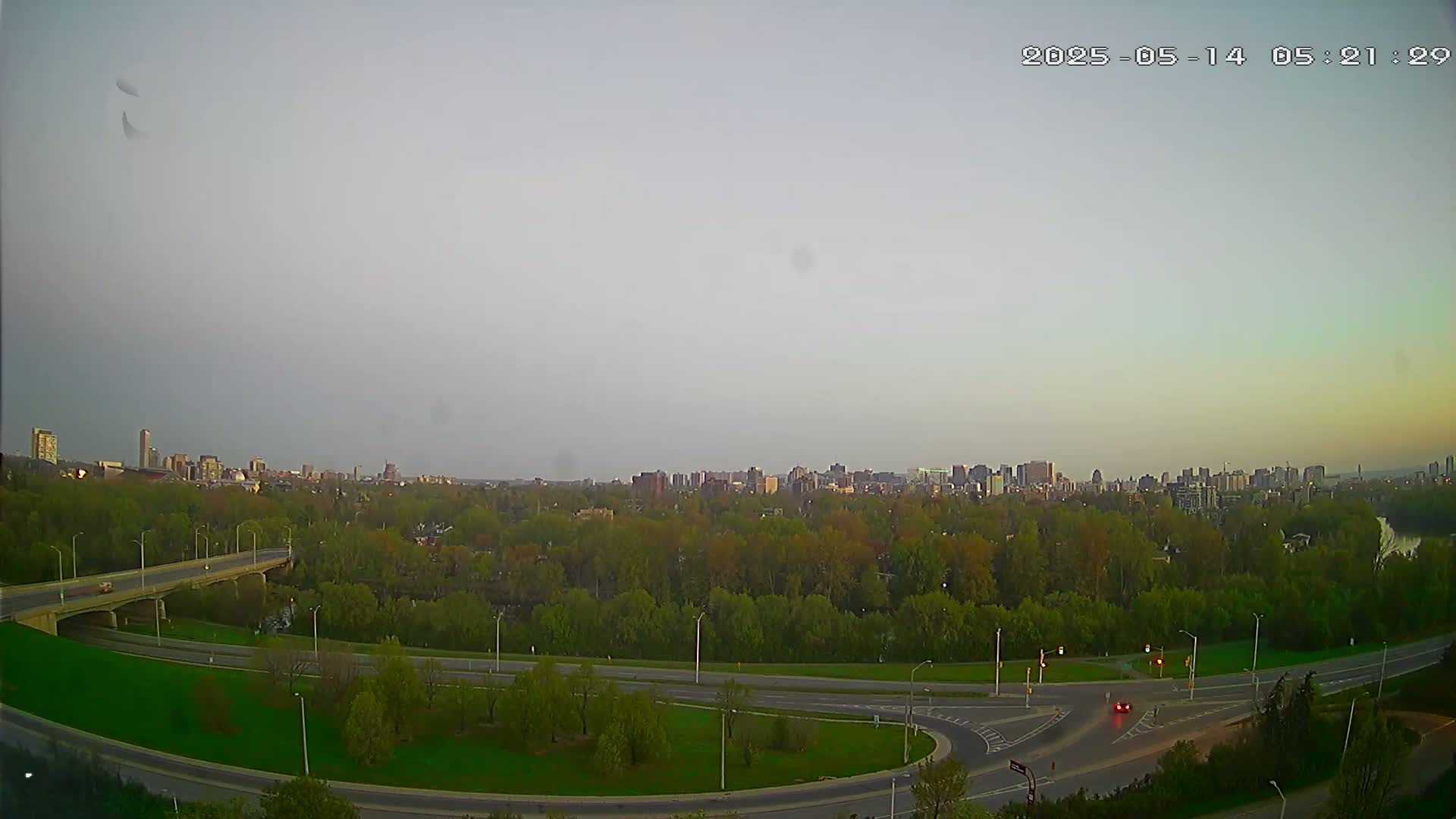 A city skyline is visible in the distance across a wide expanse of green trees and a highway interchange under a pale, overcast sky.