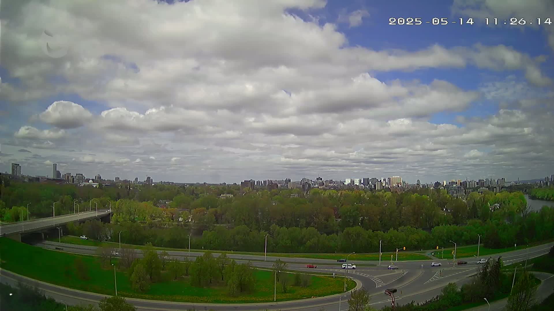 A partly cloudy sky overlooks a highway interchange and a city skyline across a green, tree-lined riverbank.