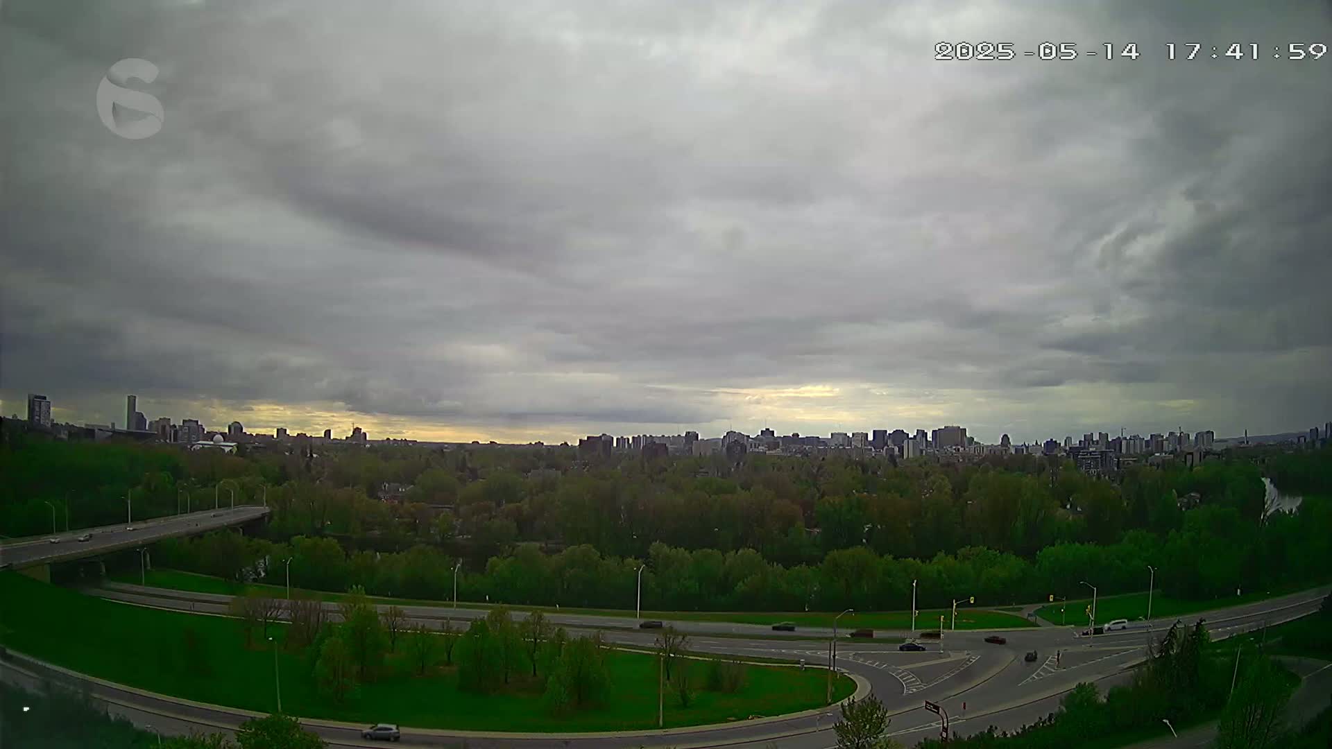 A highway curves through a lush green landscape under a cloudy sky, with a cityscape visible in the distance.